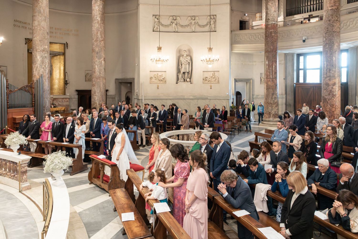Wedding guests stand in elegant white church during ceremony with bride in traditional attire.