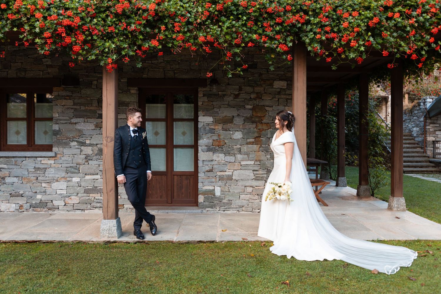 Bride and groom exchange glances outside rustic stone villa adorned with vibrant red flowering vines.