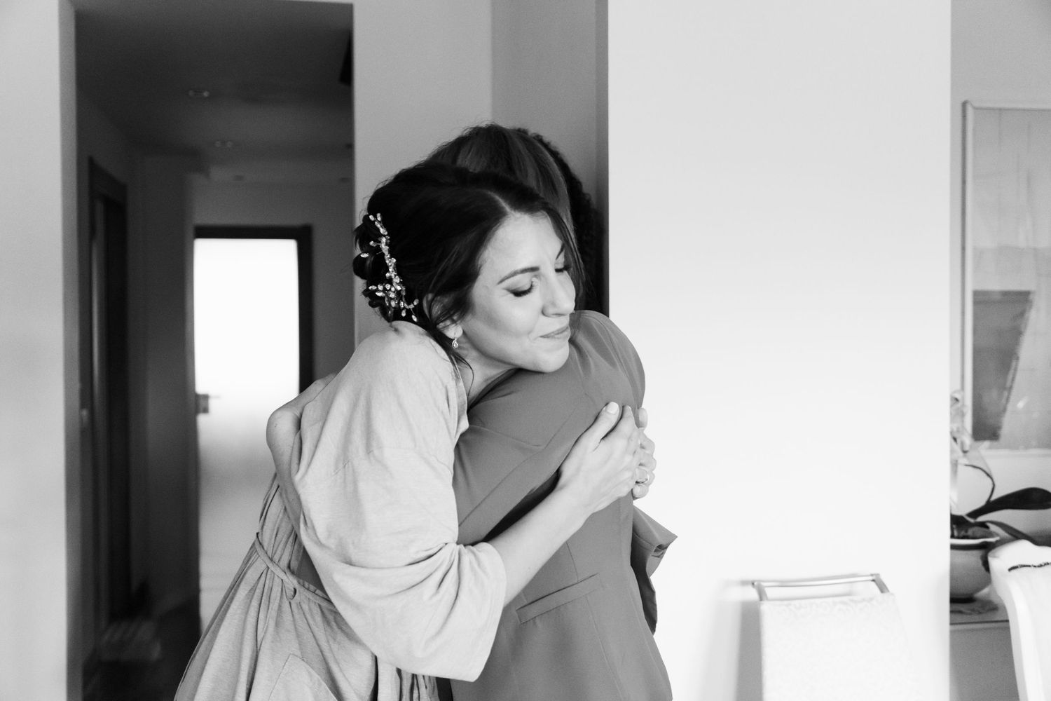 Bride in robe shares emotional hug with guest during wedding morning preparations.