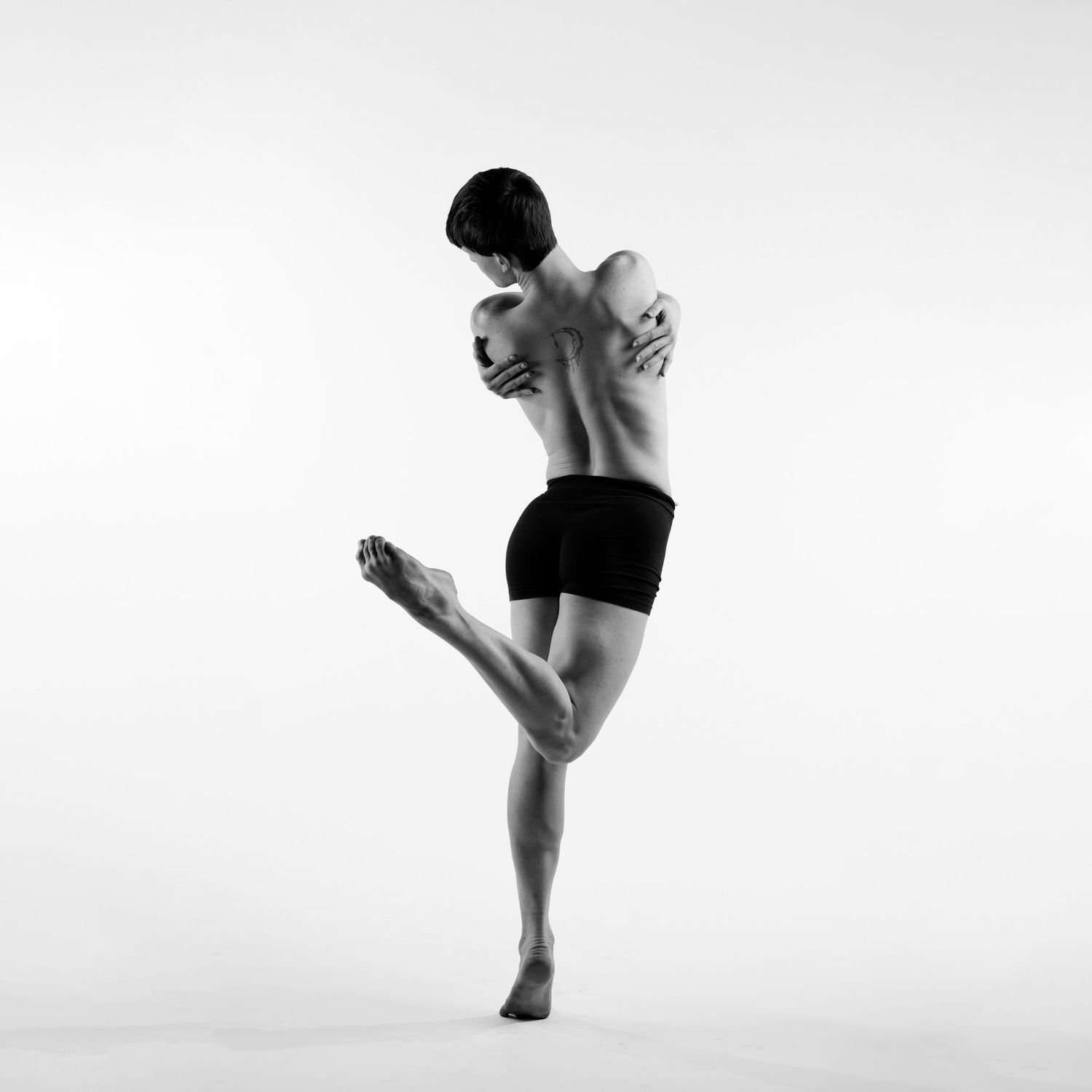 Muscular male dancer in black shorts balancing on one leg with arms crossed behind back.