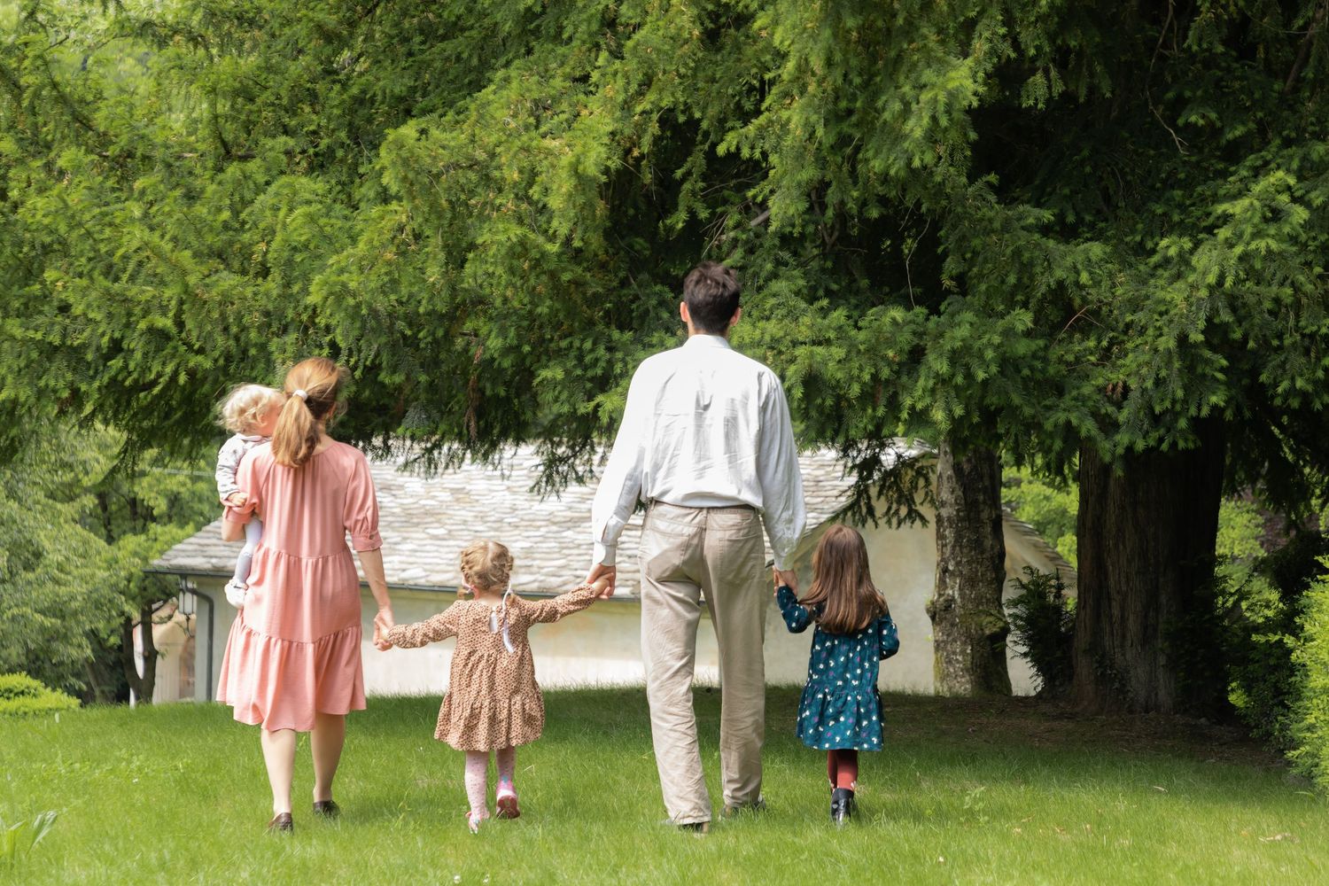 Family of five walking hand in hand across green lawn toward trees.