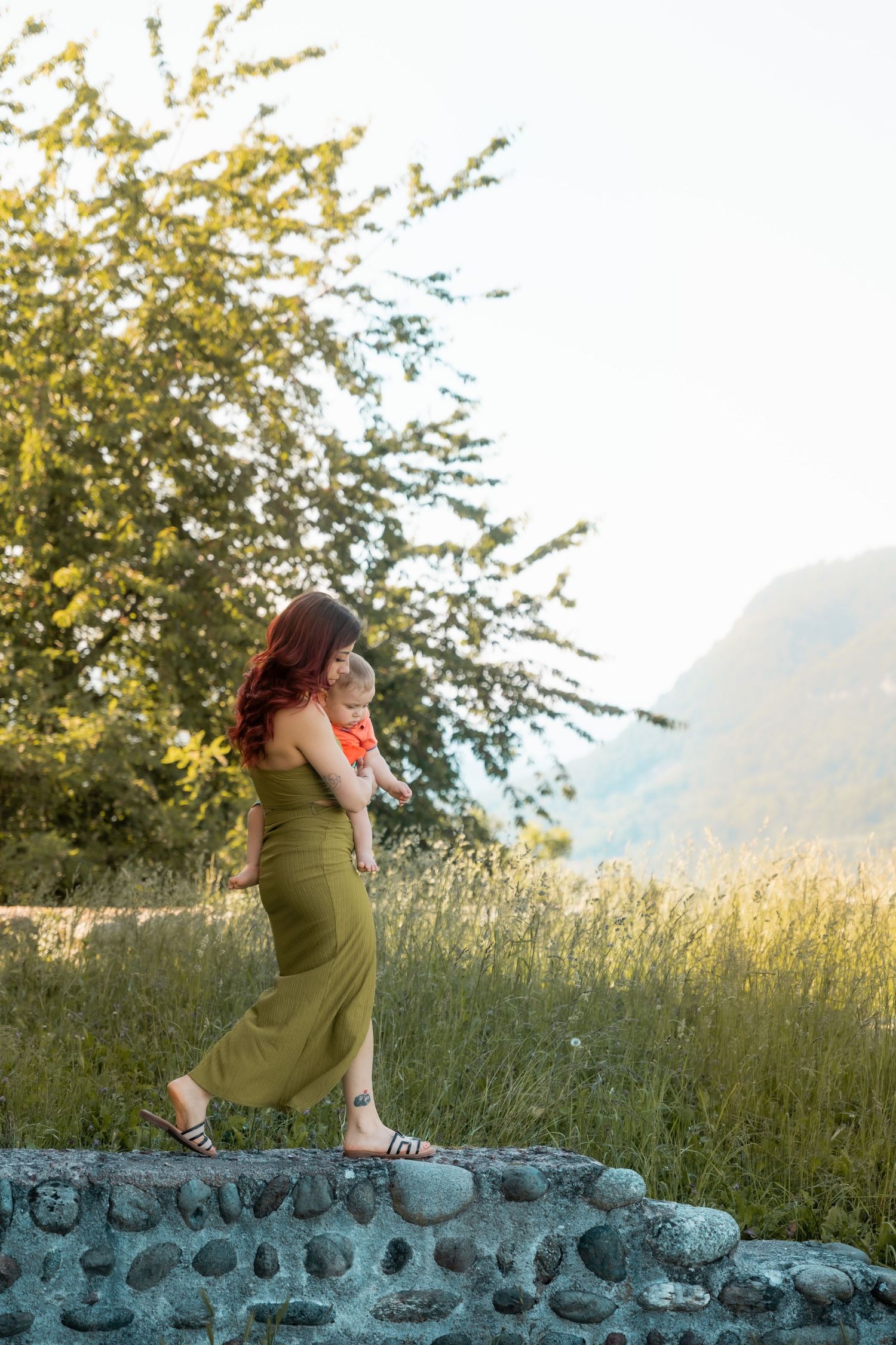 Red-haired mother in olive dress holding baby while standing on stone wall with mountain landscape behind her.
