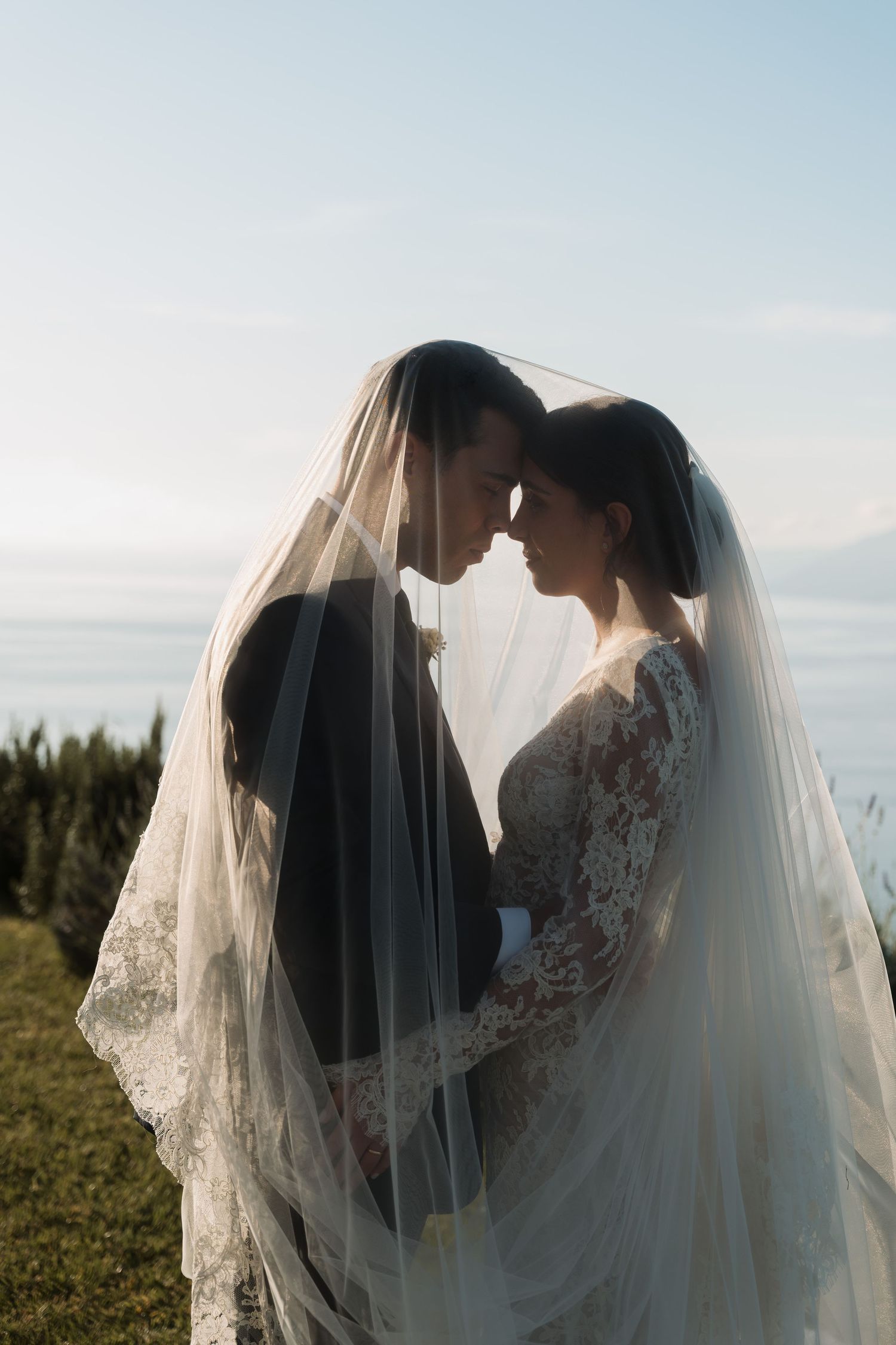 Bride and groom sharing intimate moment under flowing veil with ocean backdrop.