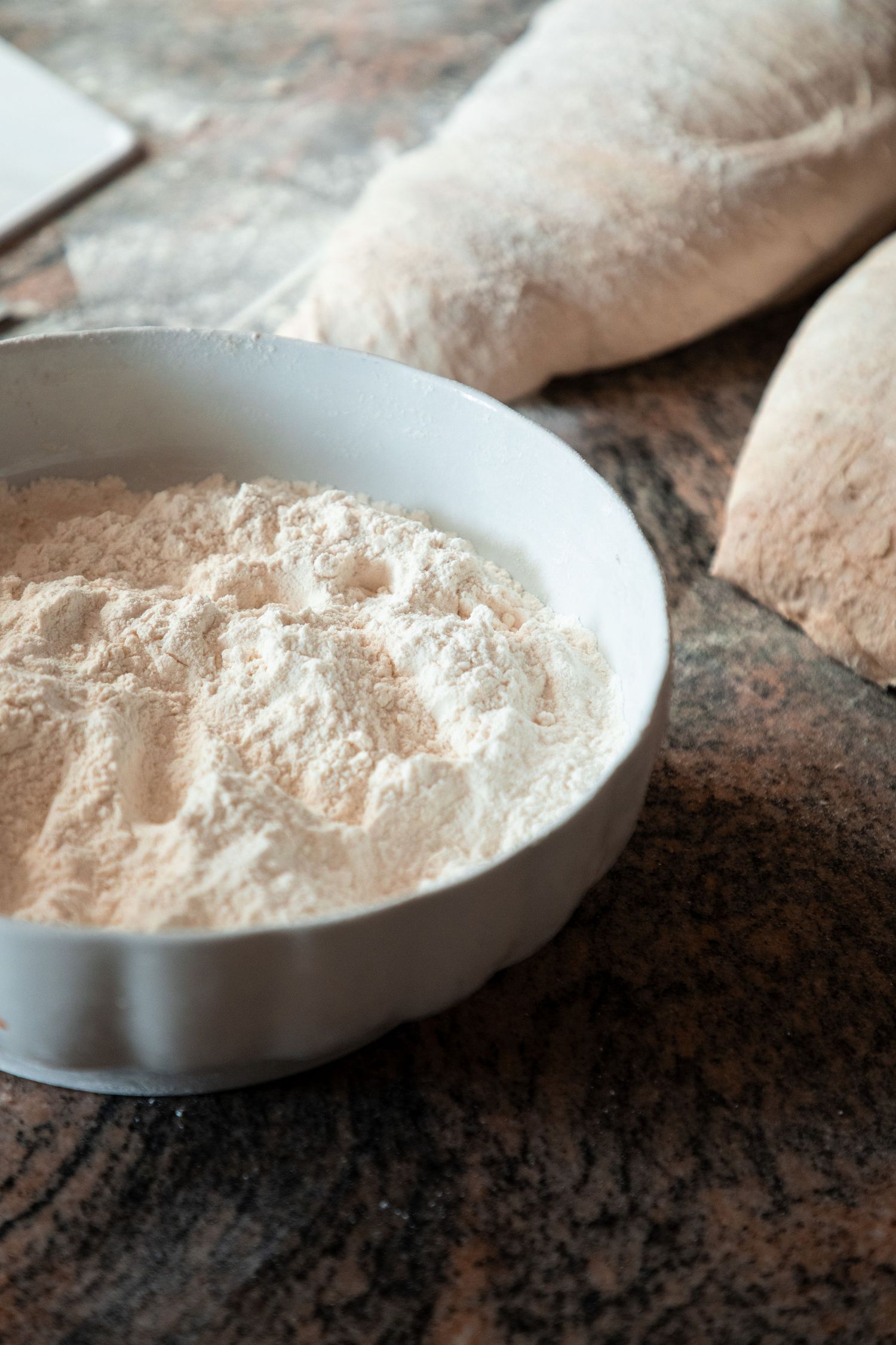 Bowl of whole wheat flour on marble surface with shaped bread dough loaves in background.