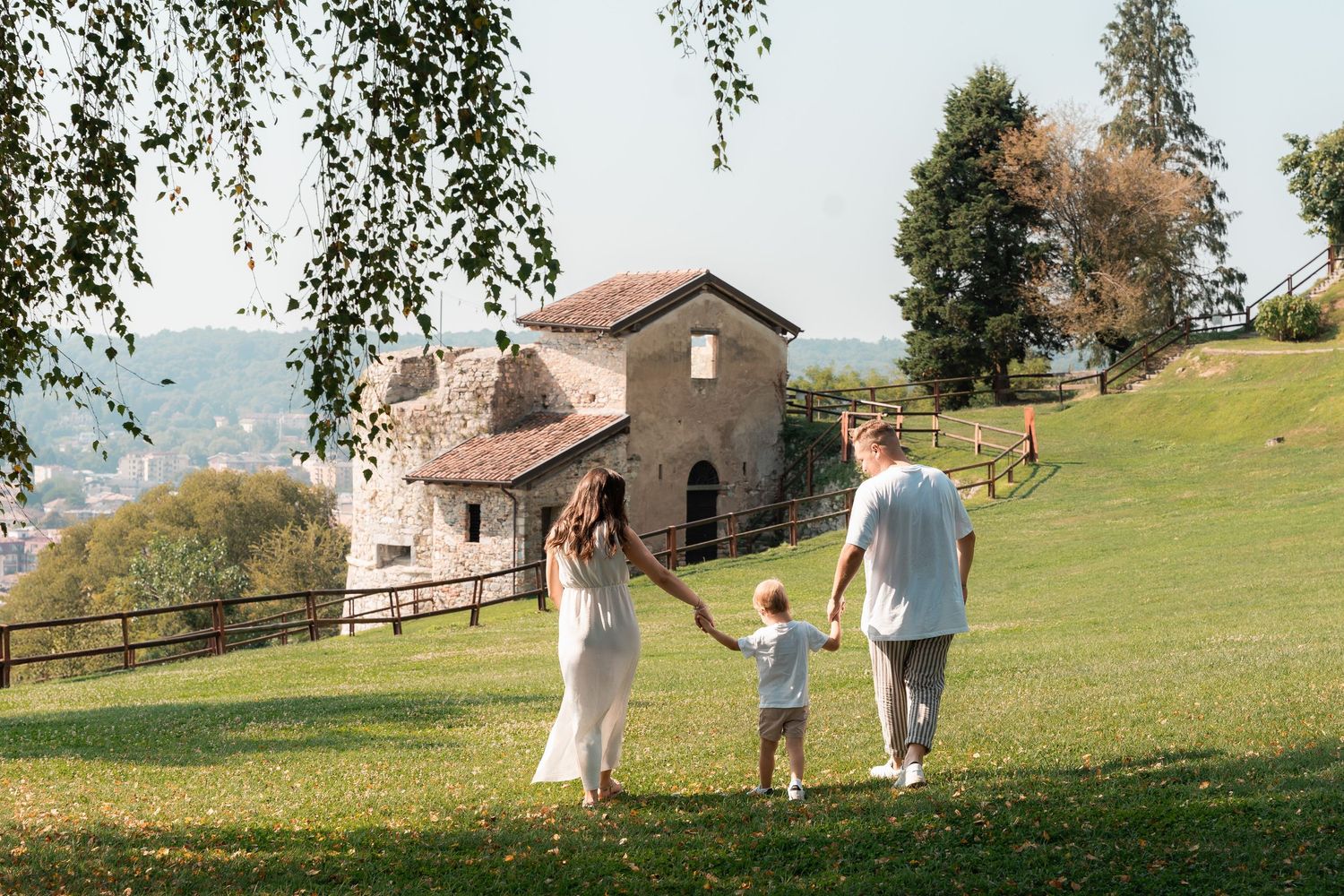 A young family holding hands walking toward a rustic stone chapel on a sunny green hillside.