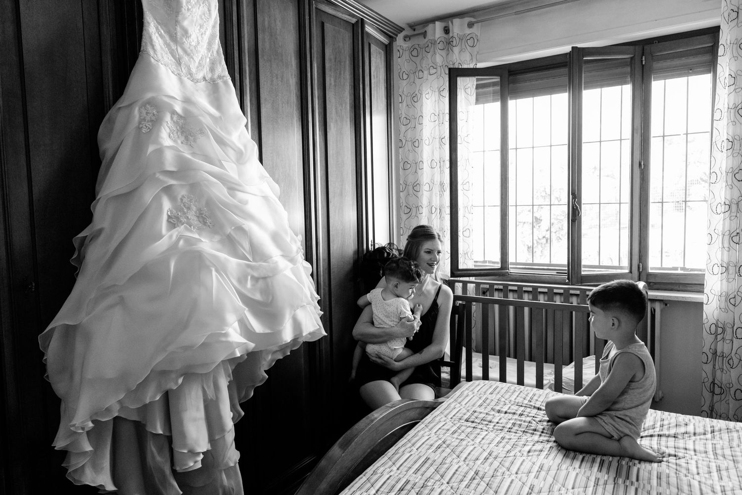 Bride in robe sits with children on bed beside hanging wedding dress before ceremony.
