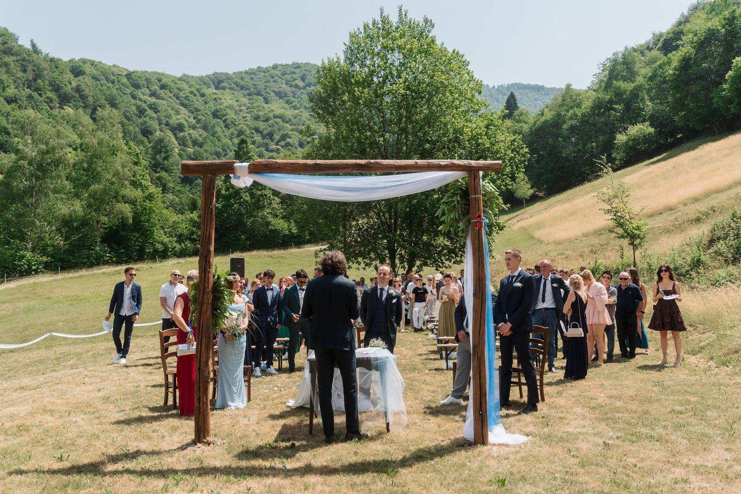 Outdoor wedding ceremony under wooden arch with white draping set against beautiful green hillside landscape.
