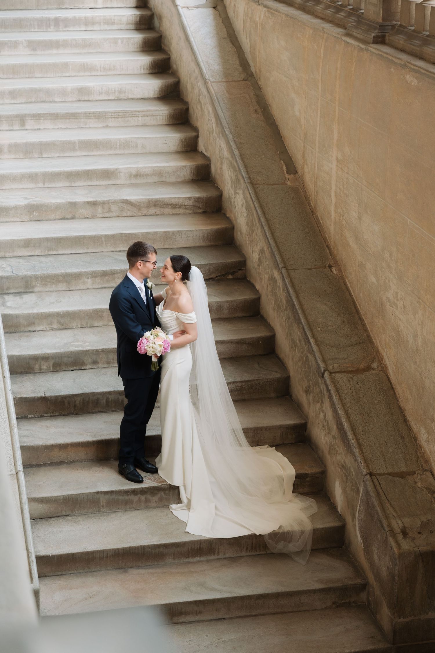 Bride and groom share a romantic embrace on grand marble staircase during wedding portrait session.
