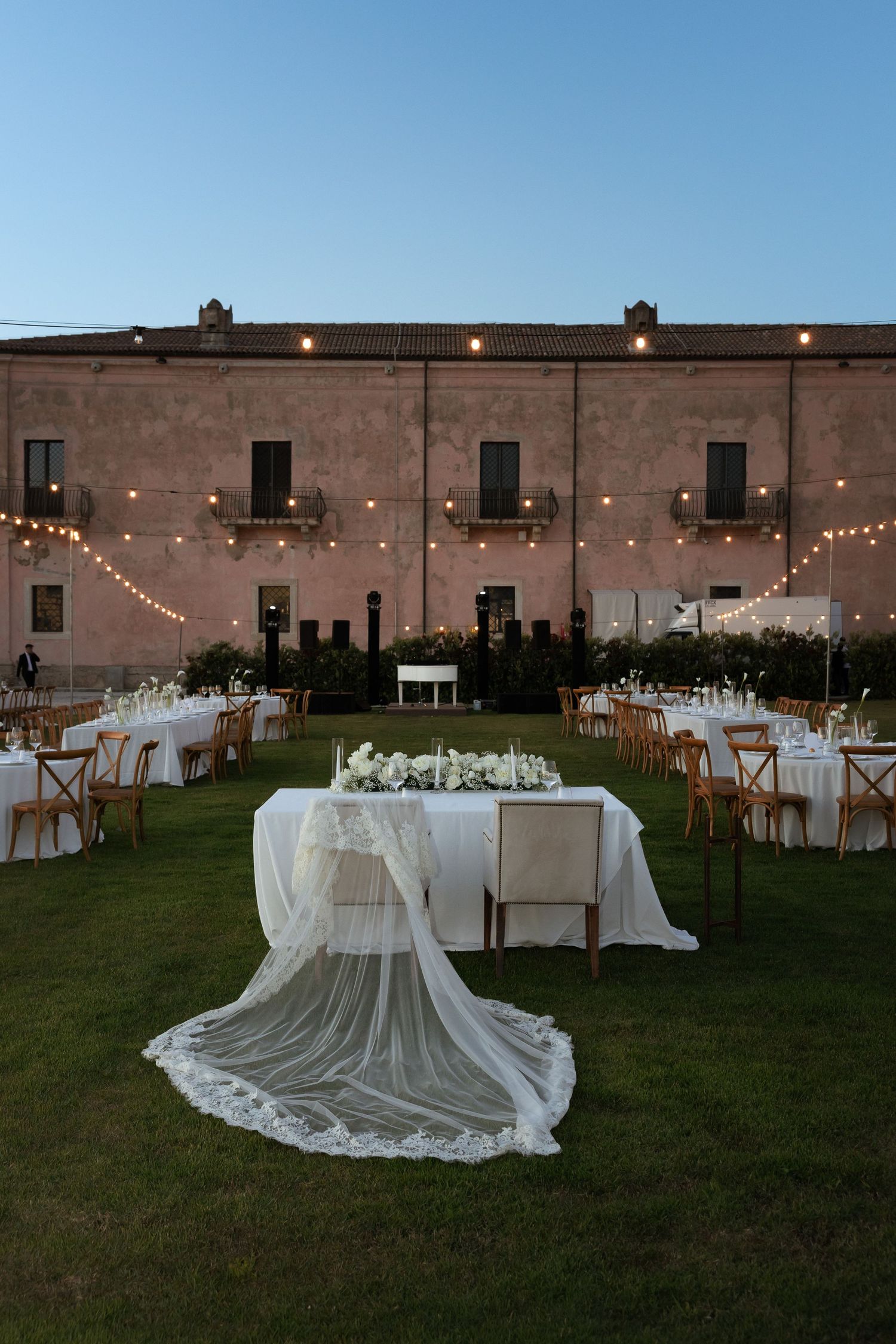 Outdoor wedding reception setup with string lights and draped sweetheart table at dusk.