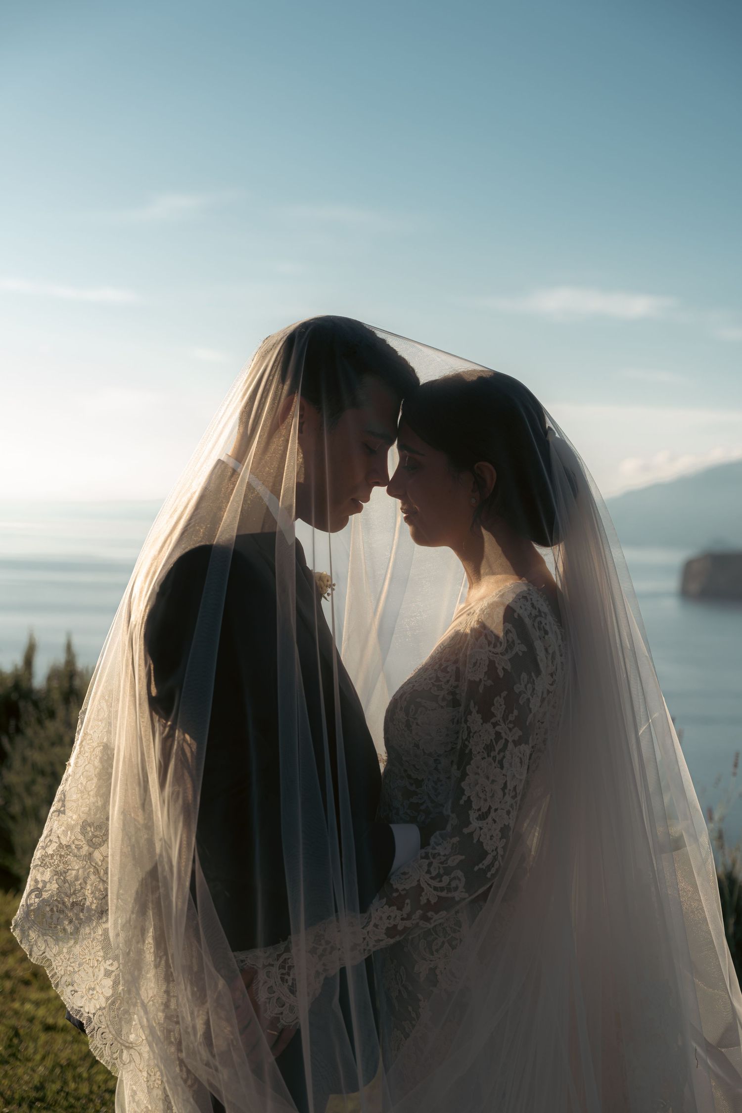 Bride and groom share an intimate moment under a wedding veil with a scenic lake and mountains in the background.