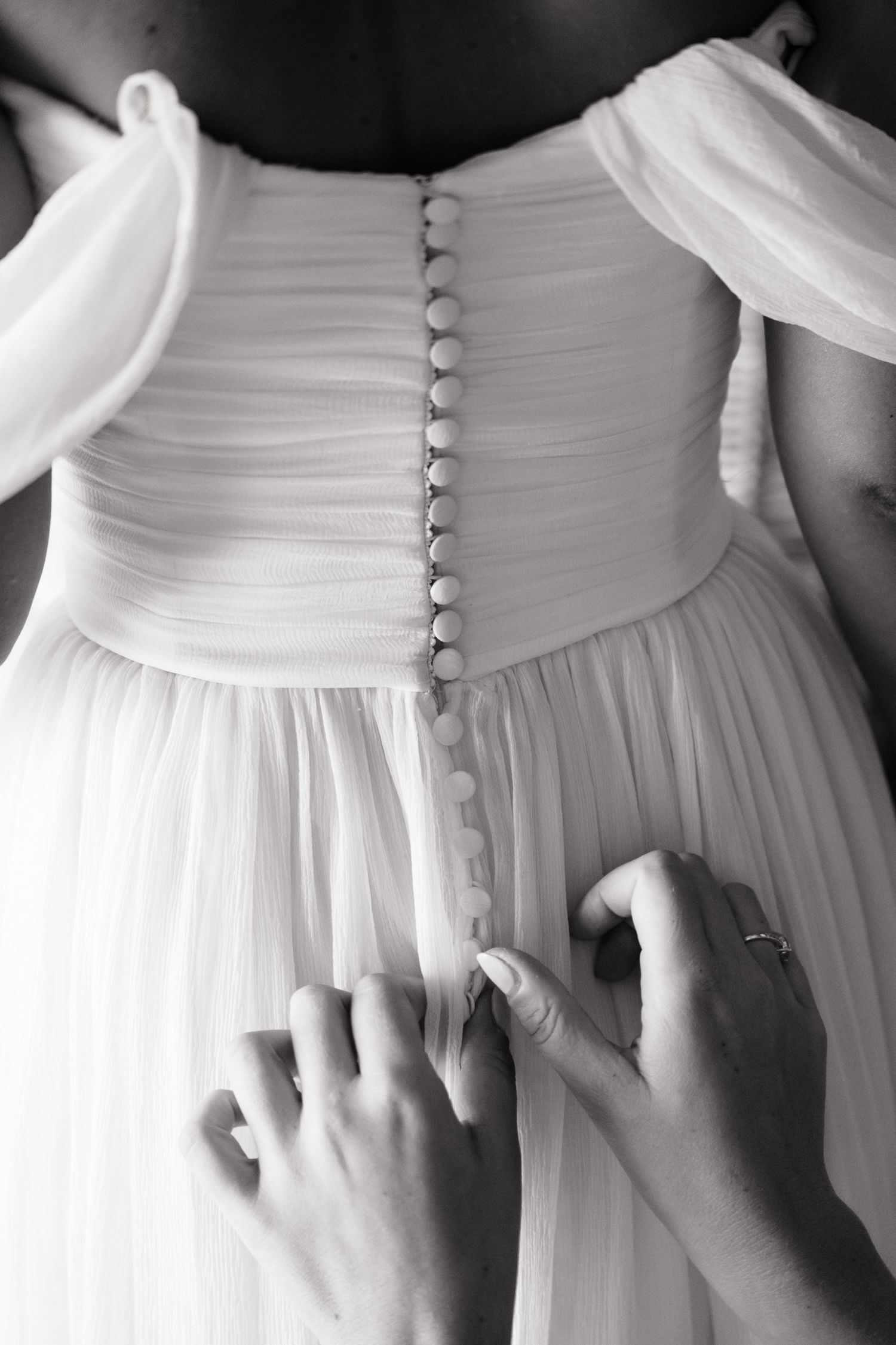 Black and white photo of hands buttoning the back of a white wedding dress with delicate buttons.