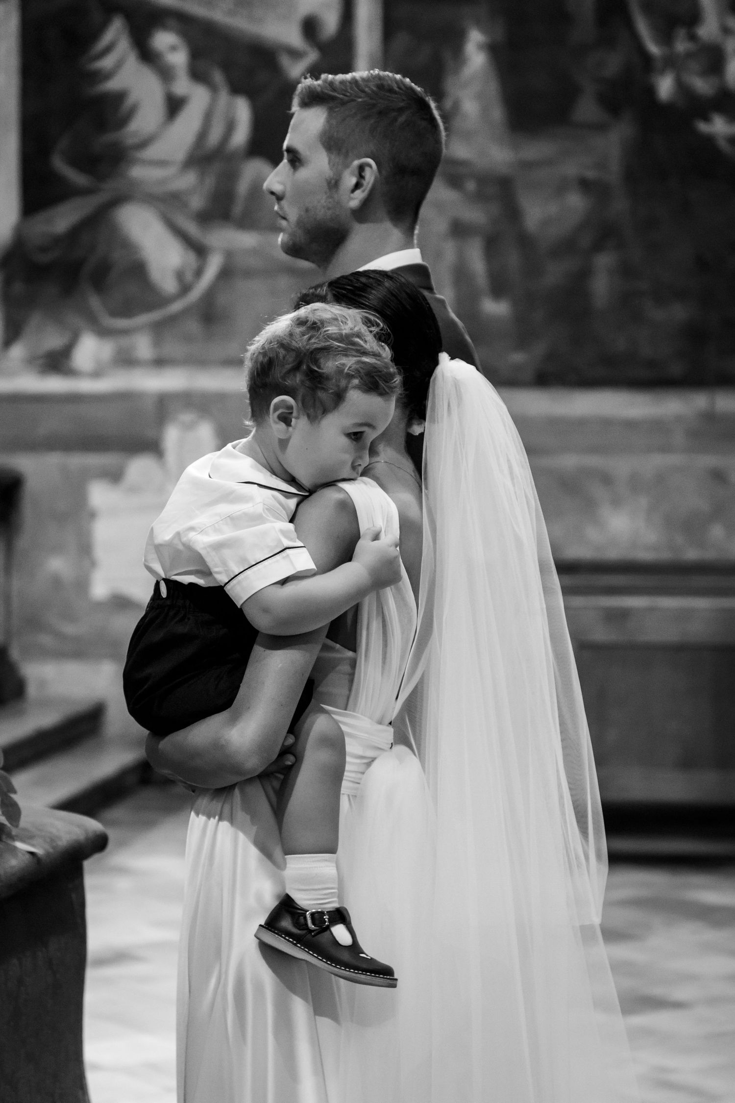 Black and white photo of bride in wedding dress holding young child during church wedding ceremony.