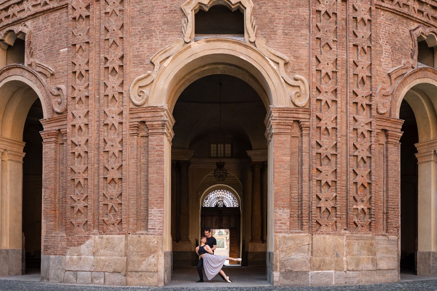 Ballet dancers share intimate moment seated in arched doorway of ornate historic brick building.