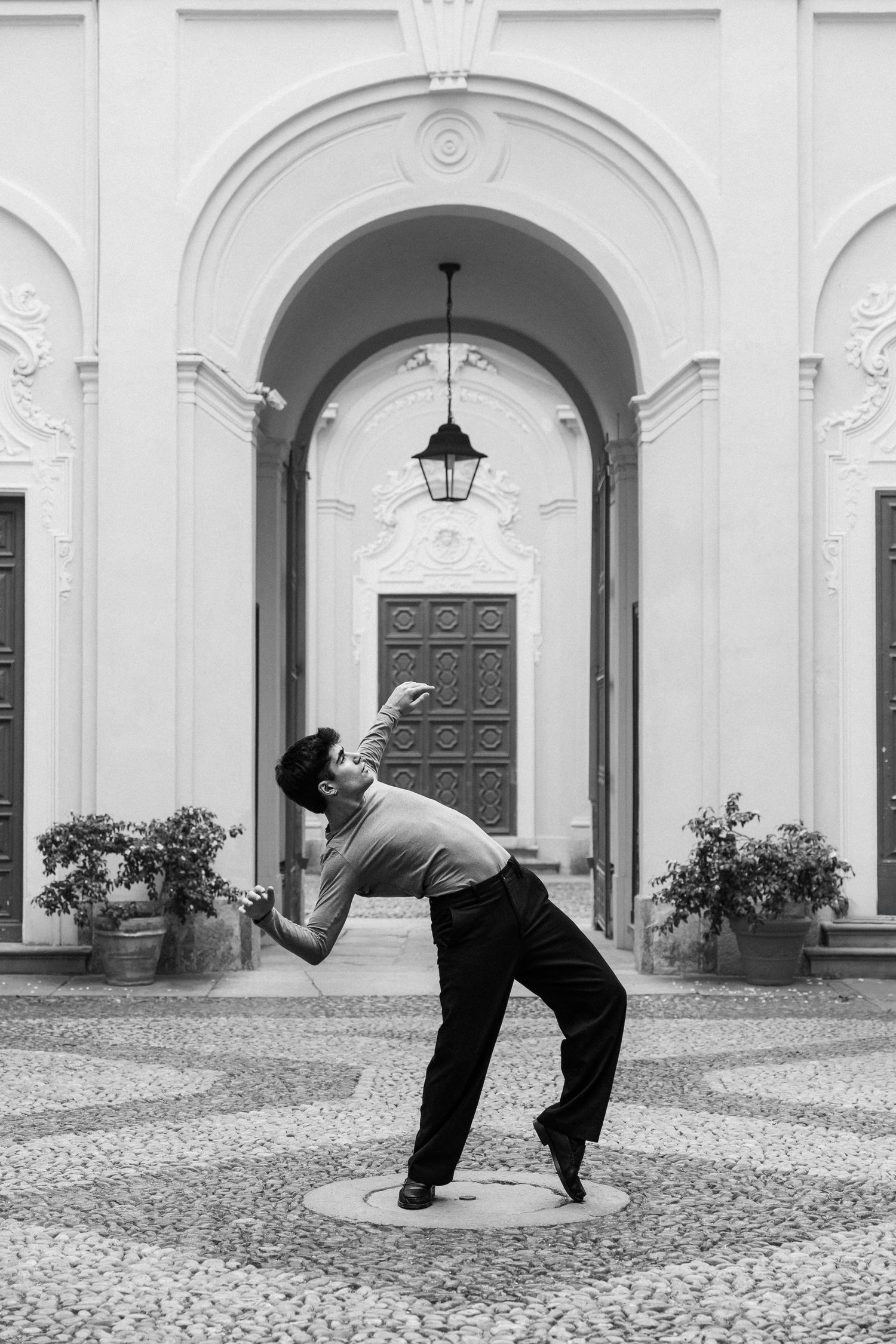 Male dancer dramatically arching backward in elegant pose before ornate classical architectural archway on cobblestone courtyard.