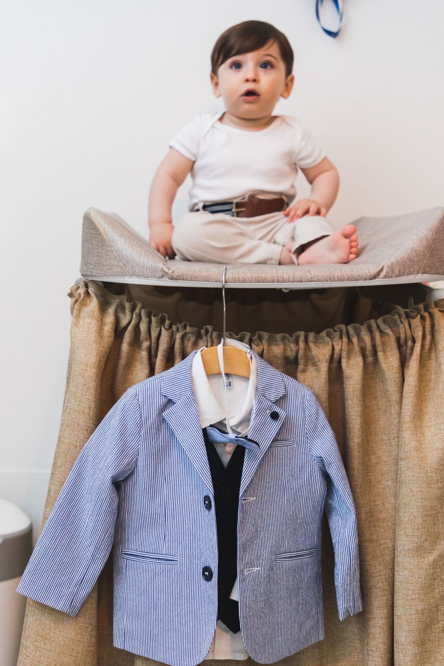 Baby boy in white onesie sitting on changing table with blue striped blazer on hanger below.
