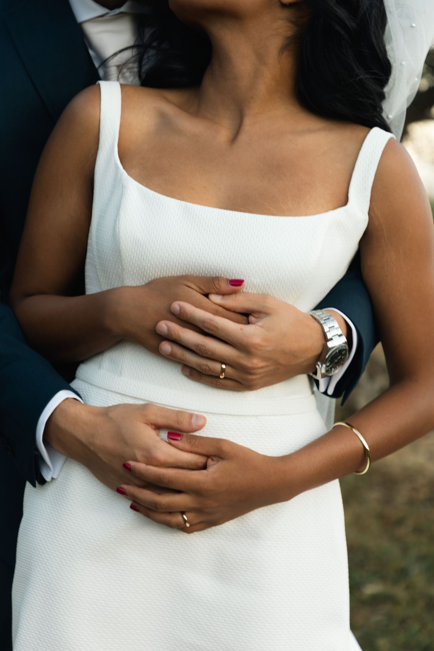 Close-up of groom embracing bride from behind, both wearing wedding rings, bride in sleek white dress.