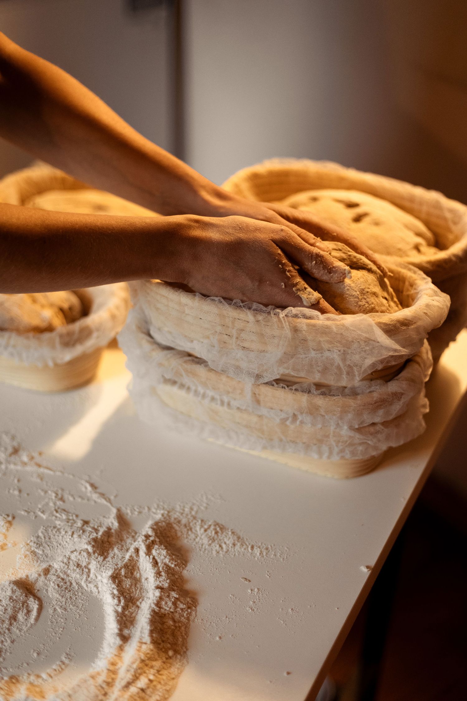 Baker's hands shaping dough in linen-lined proofing baskets dusted with flour on white surface.