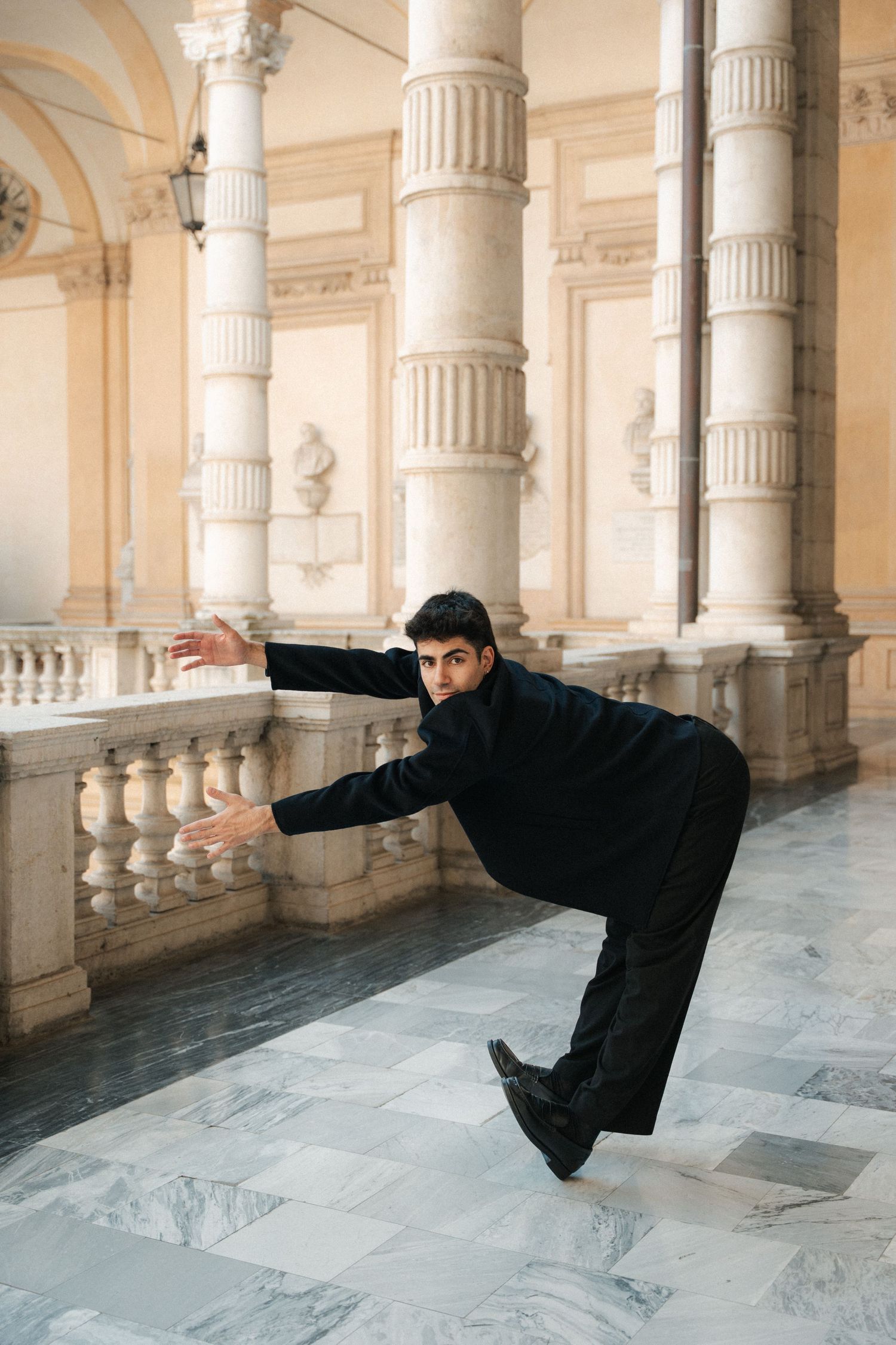 Male dancer in black leaning dramatically over marble balustrade in ornate classical colonnade.