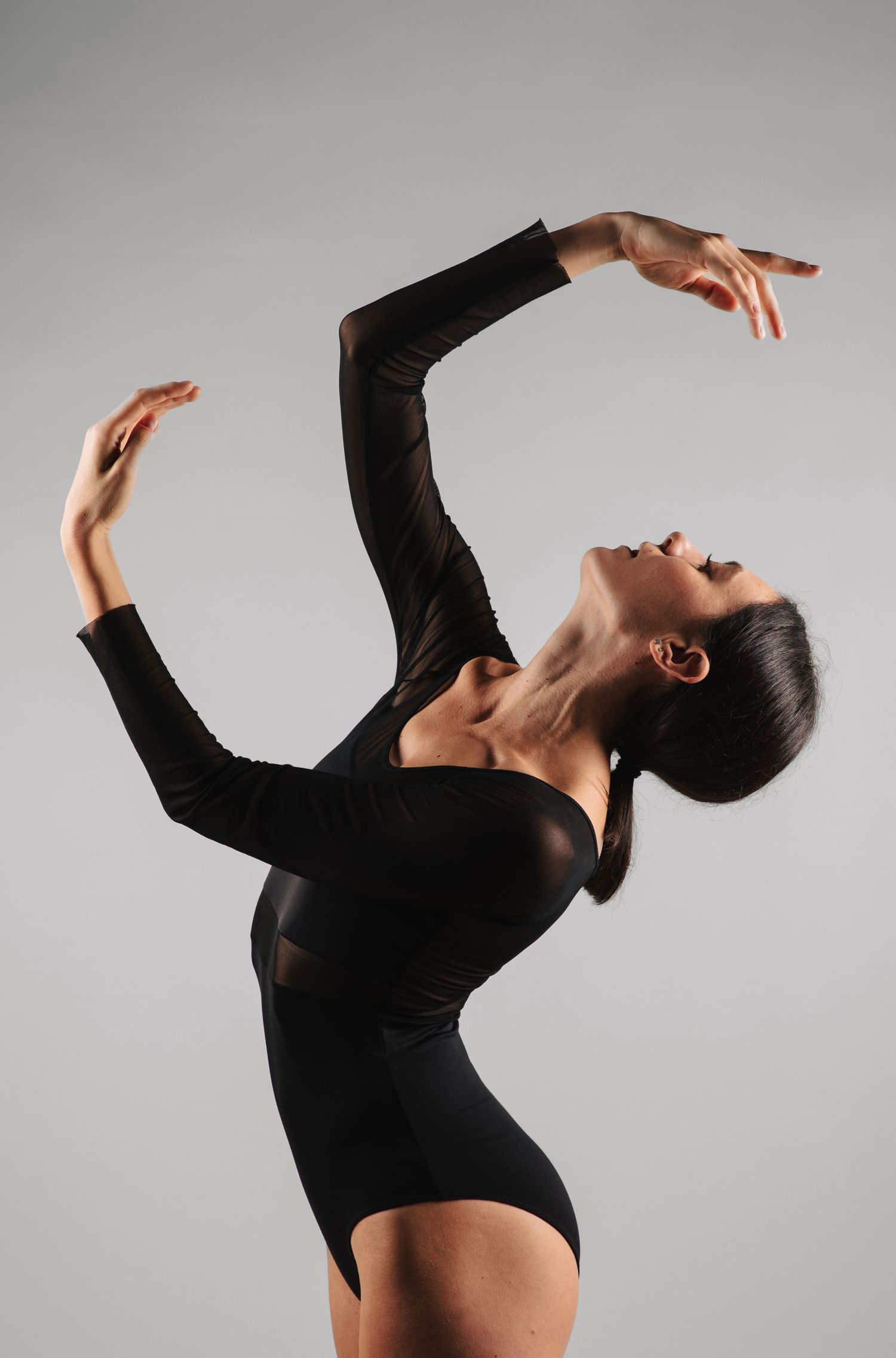 Elegant female dancer in black leotard arches back with arms raised gracefully in studio lighting.