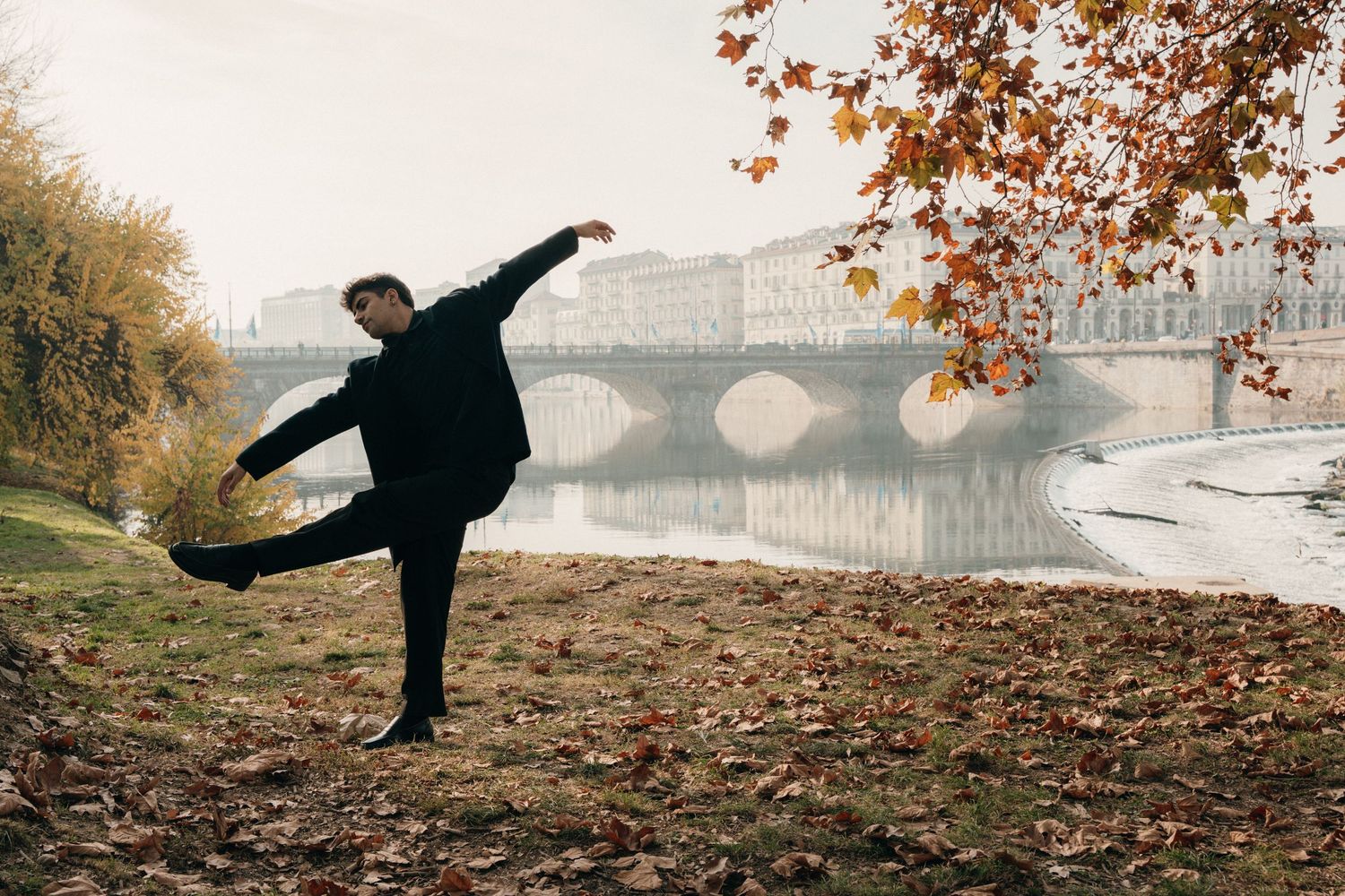 Male dancer in black balancing on one leg by autumn riverside with bridge backdrop.