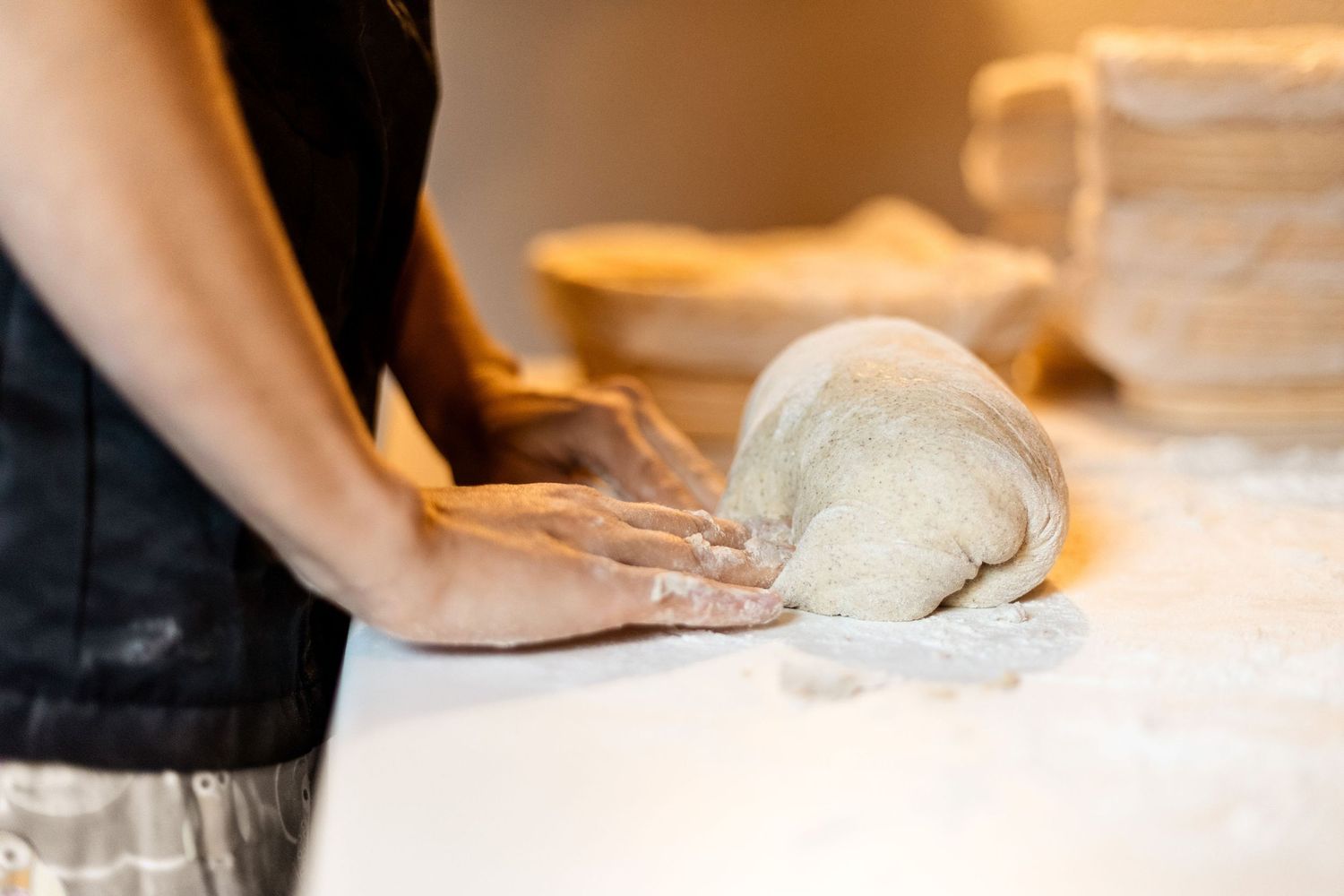 Baker kneading bread dough on flour-dusted white surface with proofing baskets in background.