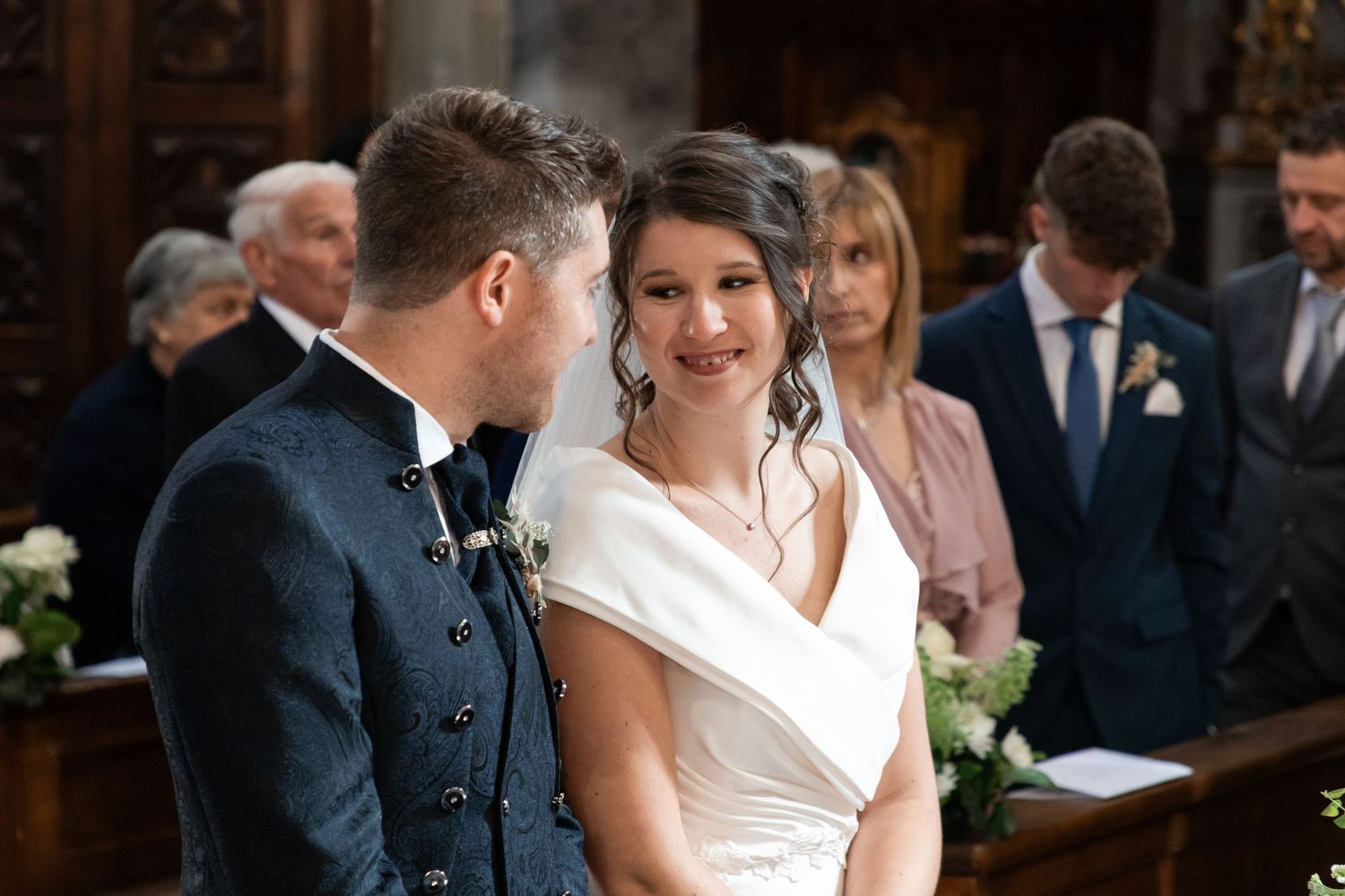 Smiling bride in white dress gazes at groom during wedding ceremony with guests seated behind them.