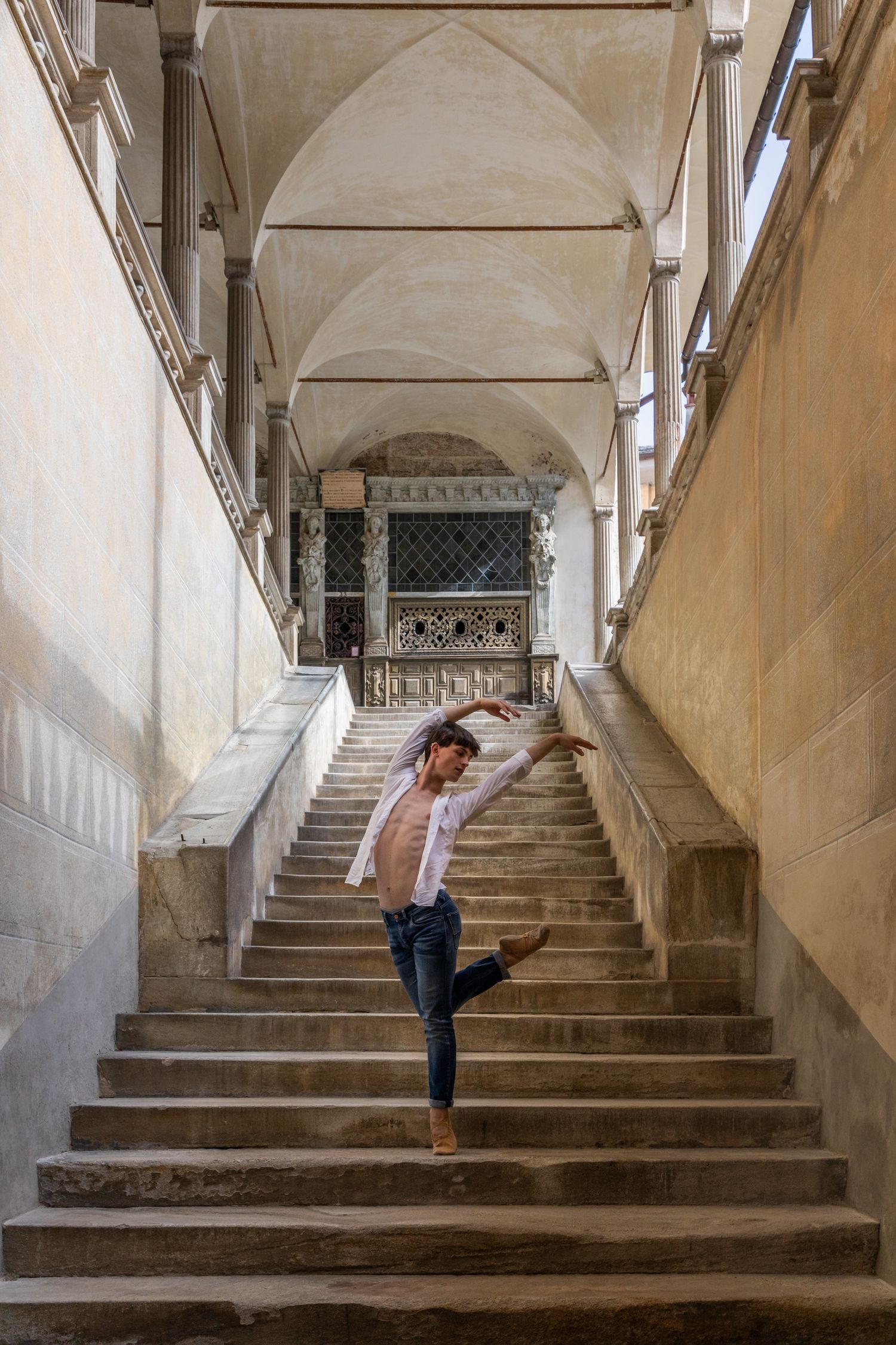 Male ballet dancer performs elegant one-legged pose on historic stone staircase with arched colonnade.