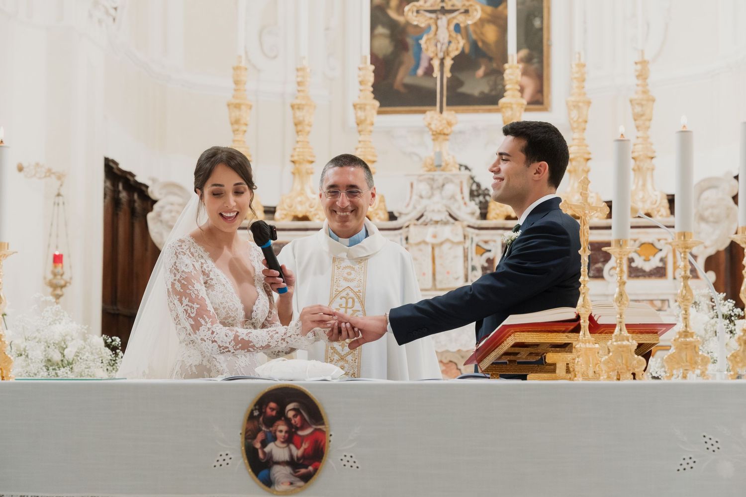 Bride and groom exchanging vows at ornate church altar with priest officiating ceremony.