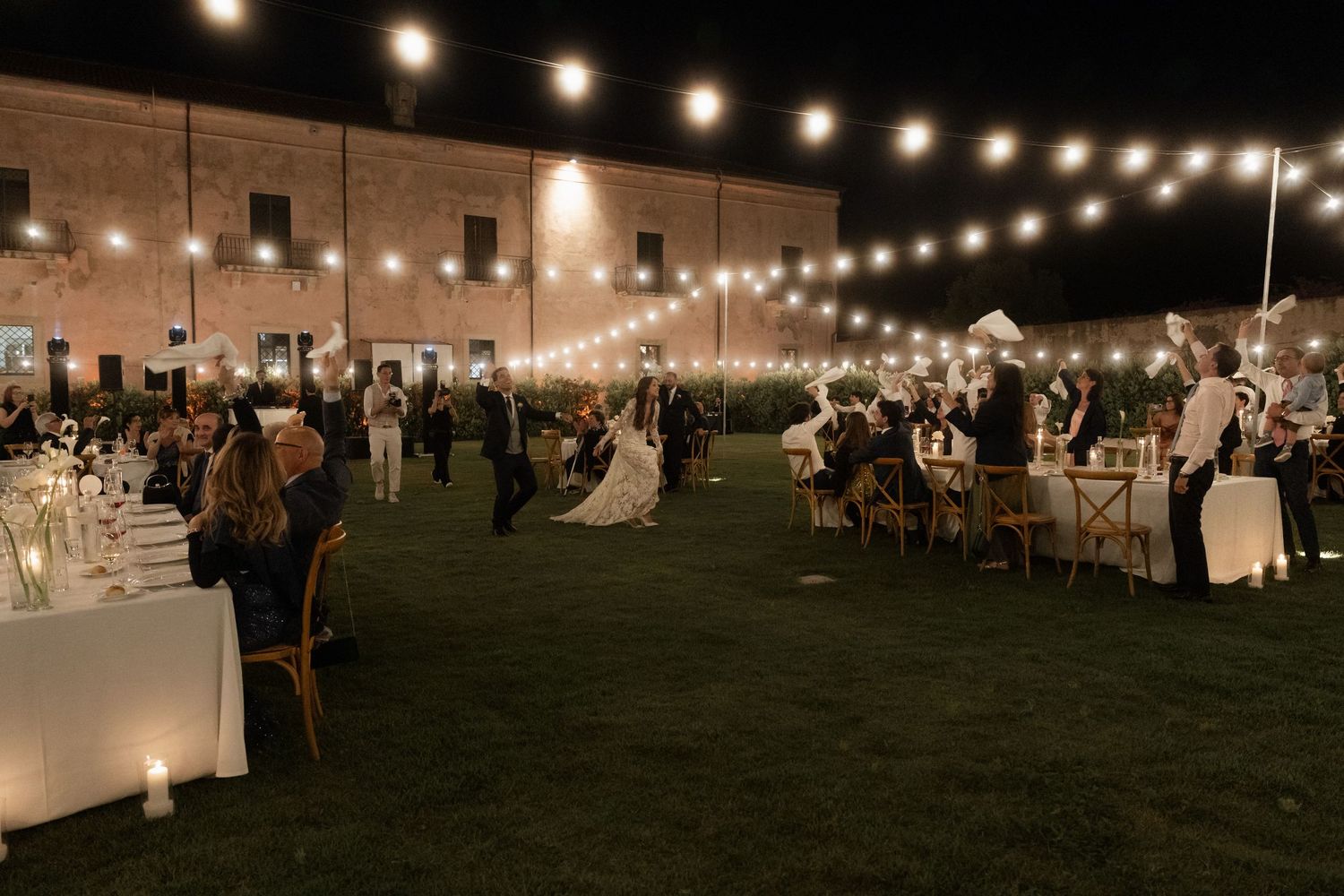 Bride and groom dancing at romantic outdoor evening reception under warm string lights.