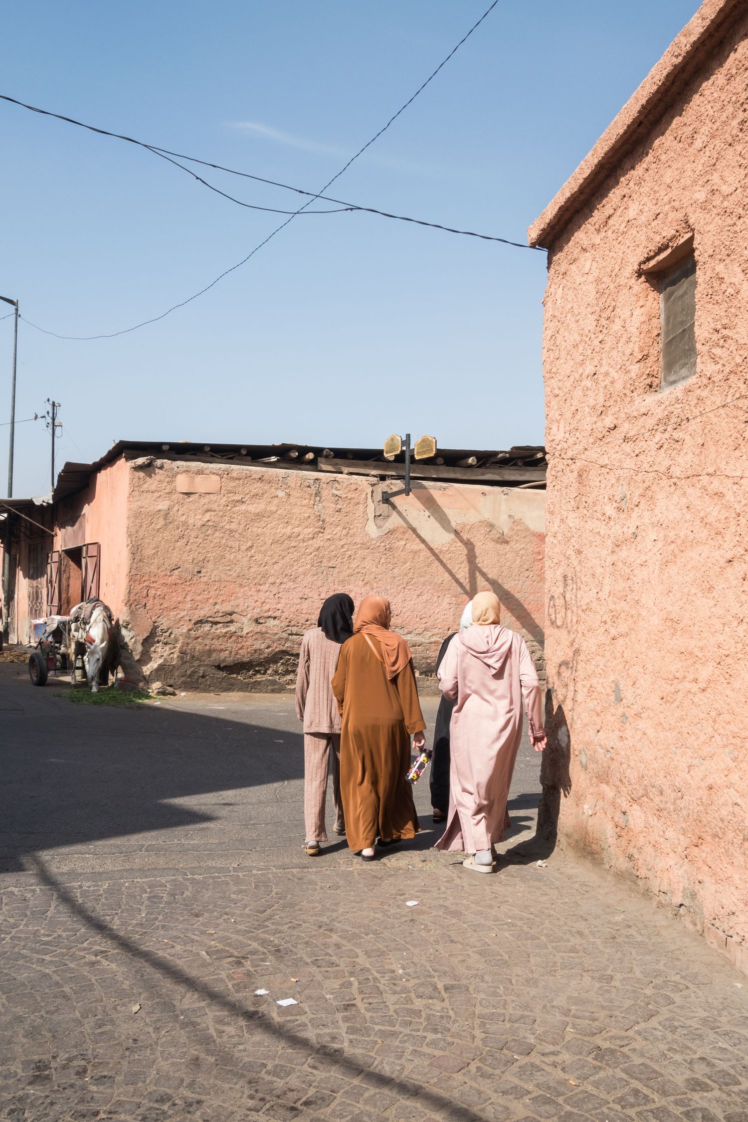 Three women in hijabs and traditional robes walking along a narrow Moroccan medina street.