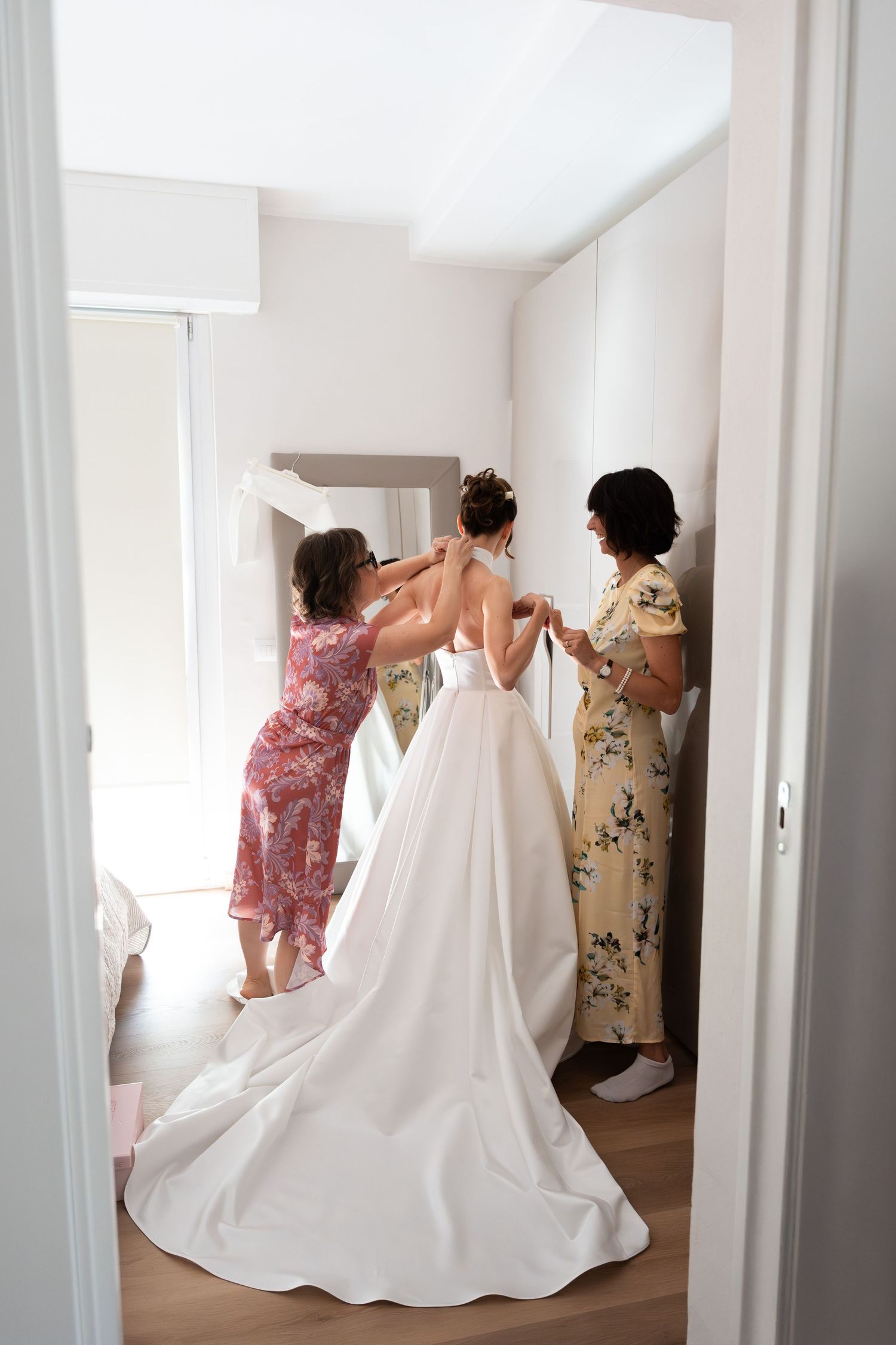 Two women in floral dresses help bride fasten her white wedding gown in a bright dressing room.