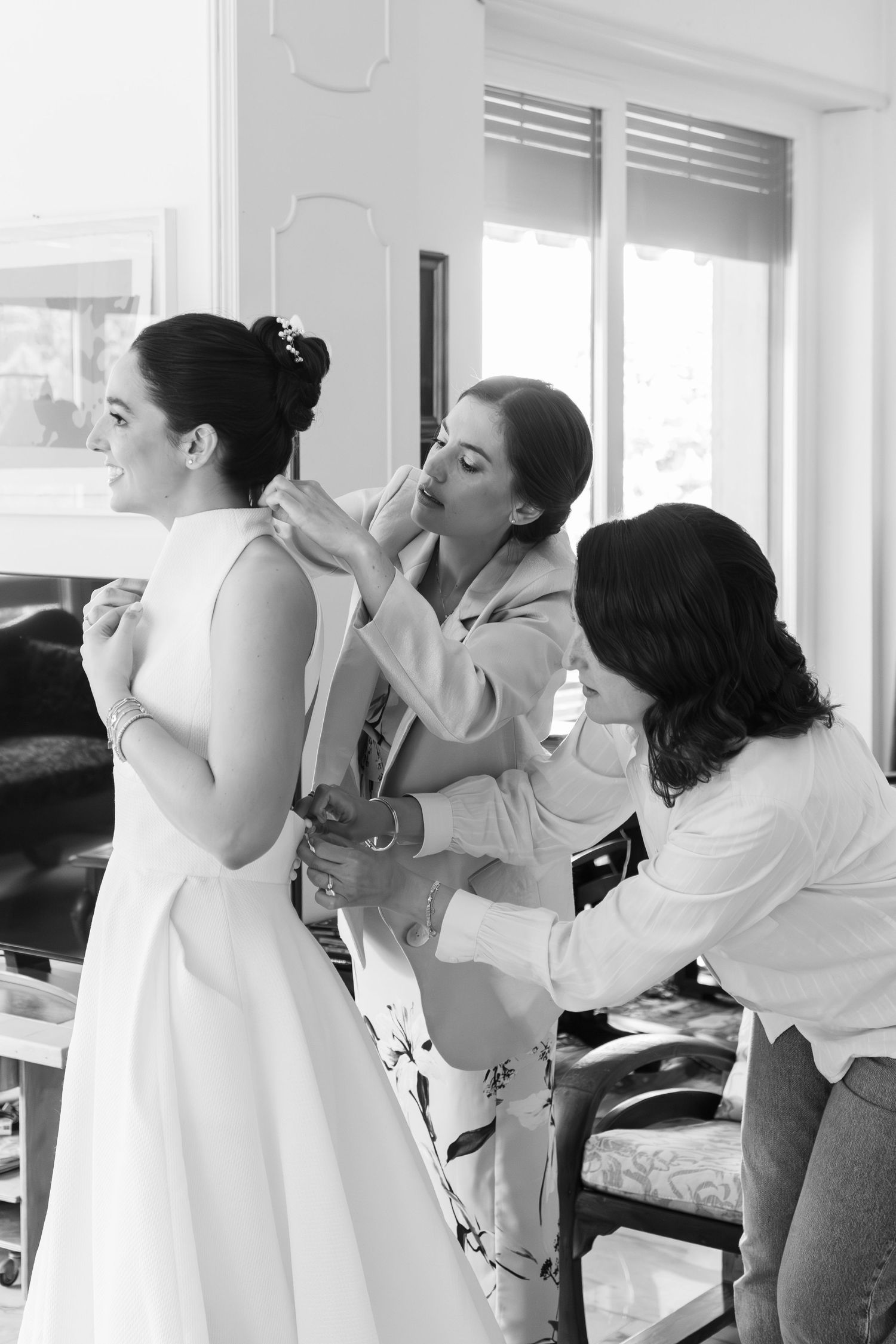 Black and white photo of two women helping smiling bride button her white wedding dress before ceremony.