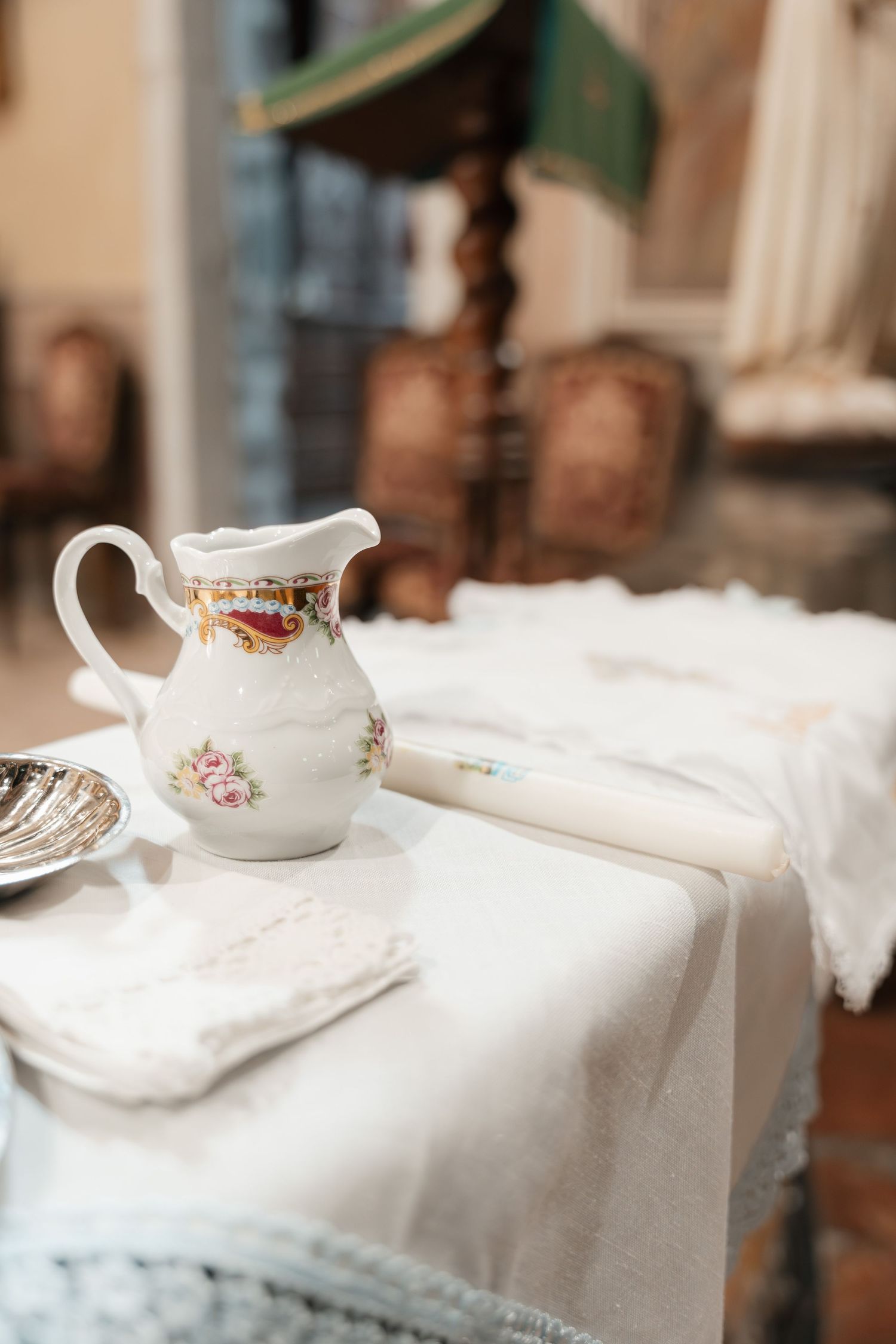 An elegant white floral porcelain pitcher on a white linen tablecloth in a vintage interior setting.