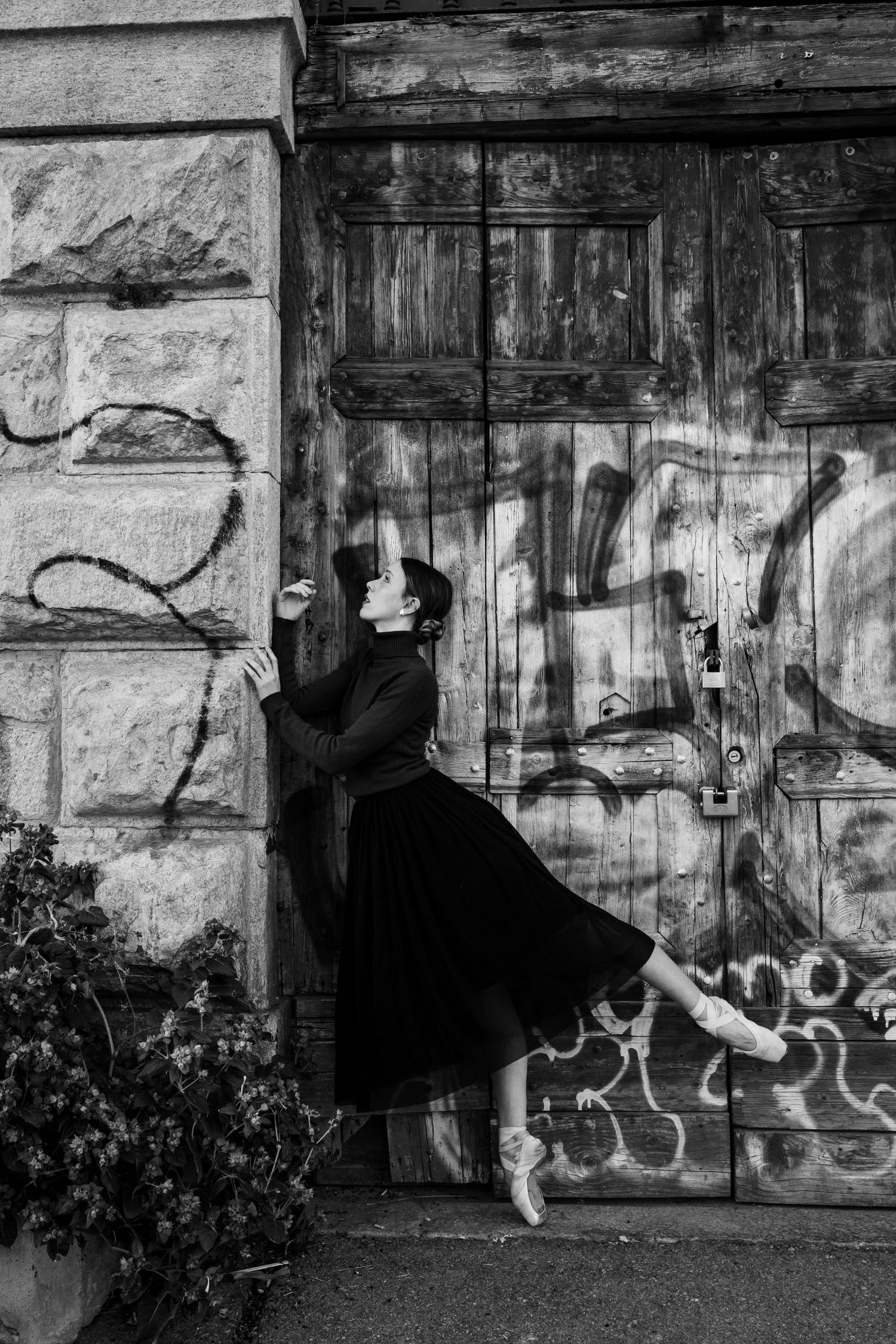 Ballerina in black dress leaping against graffiti-covered weathered wooden doors, black and white photo.