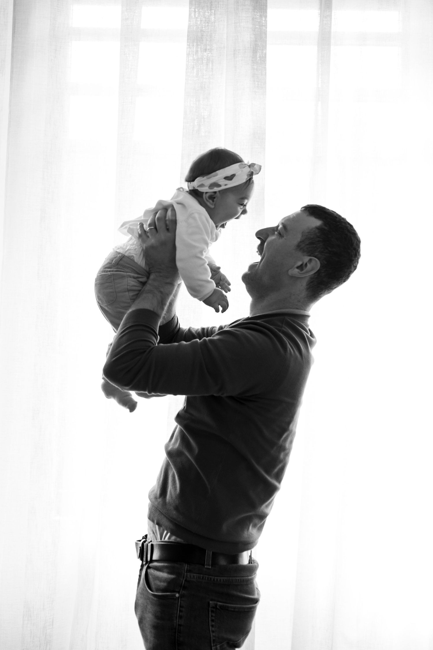 Black and white photo of father lifting smiling baby near curtained window.