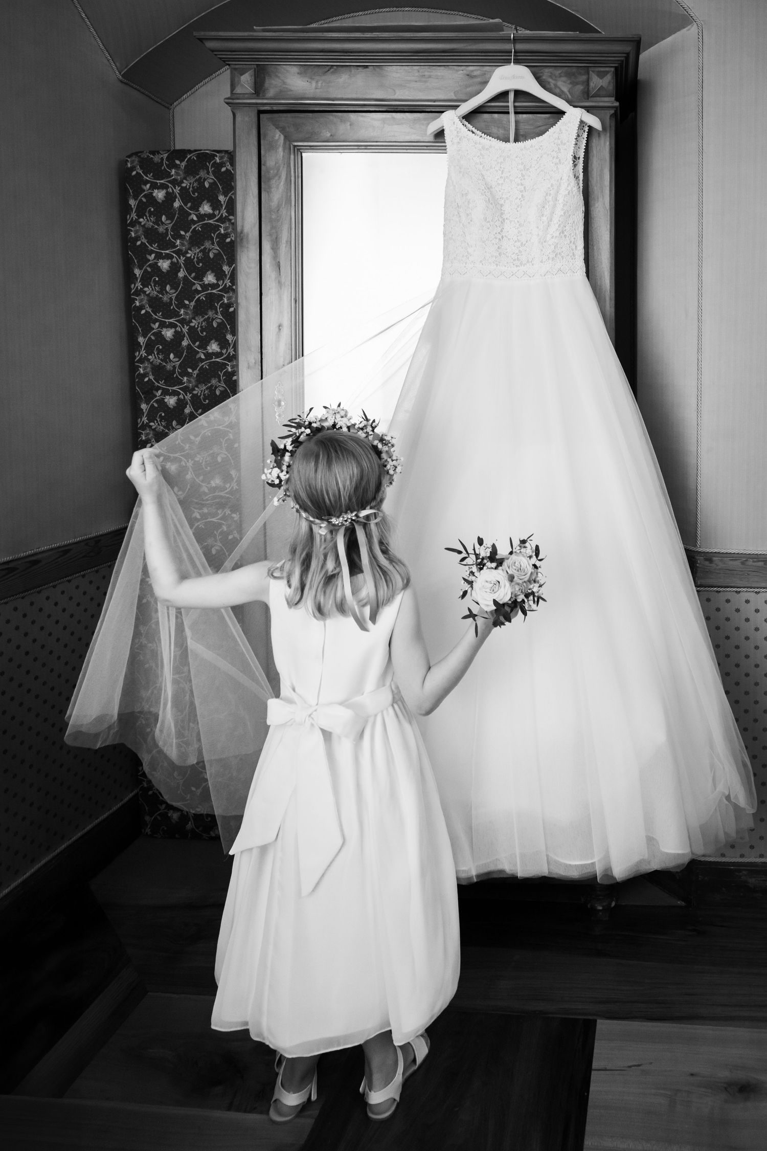 Young flower girl in white dress holds bouquet and veil before hanging wedding gown.
