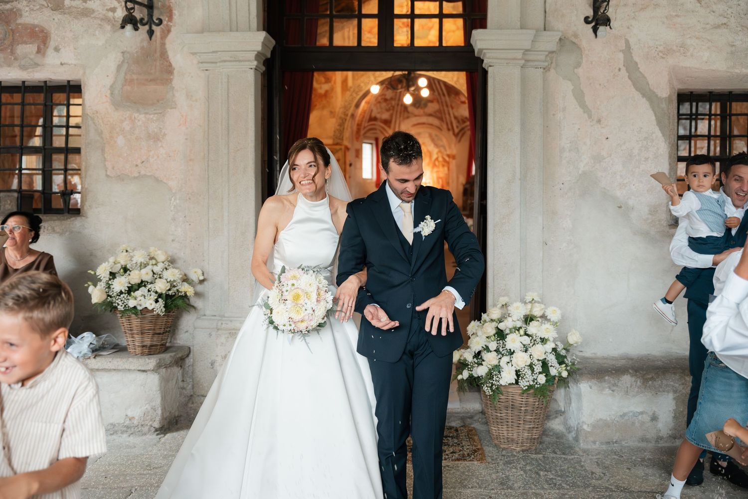 Bride in ballgown and groom exit church entrance flanked by floral arrangements and guests.