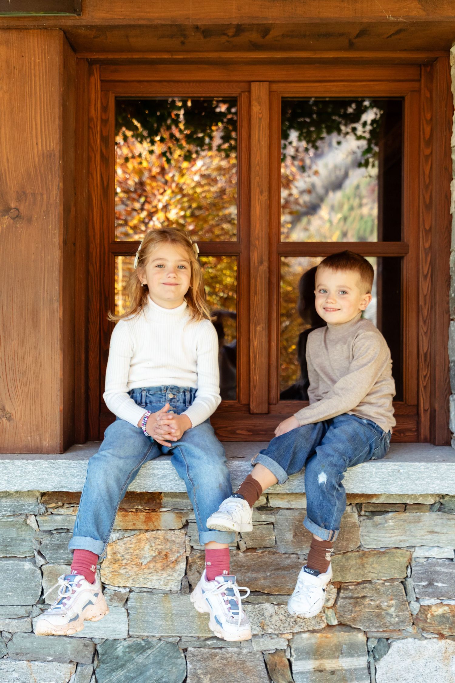 Two young children in casual clothes sit together on stone steps outside a wooden cabin in autumn.