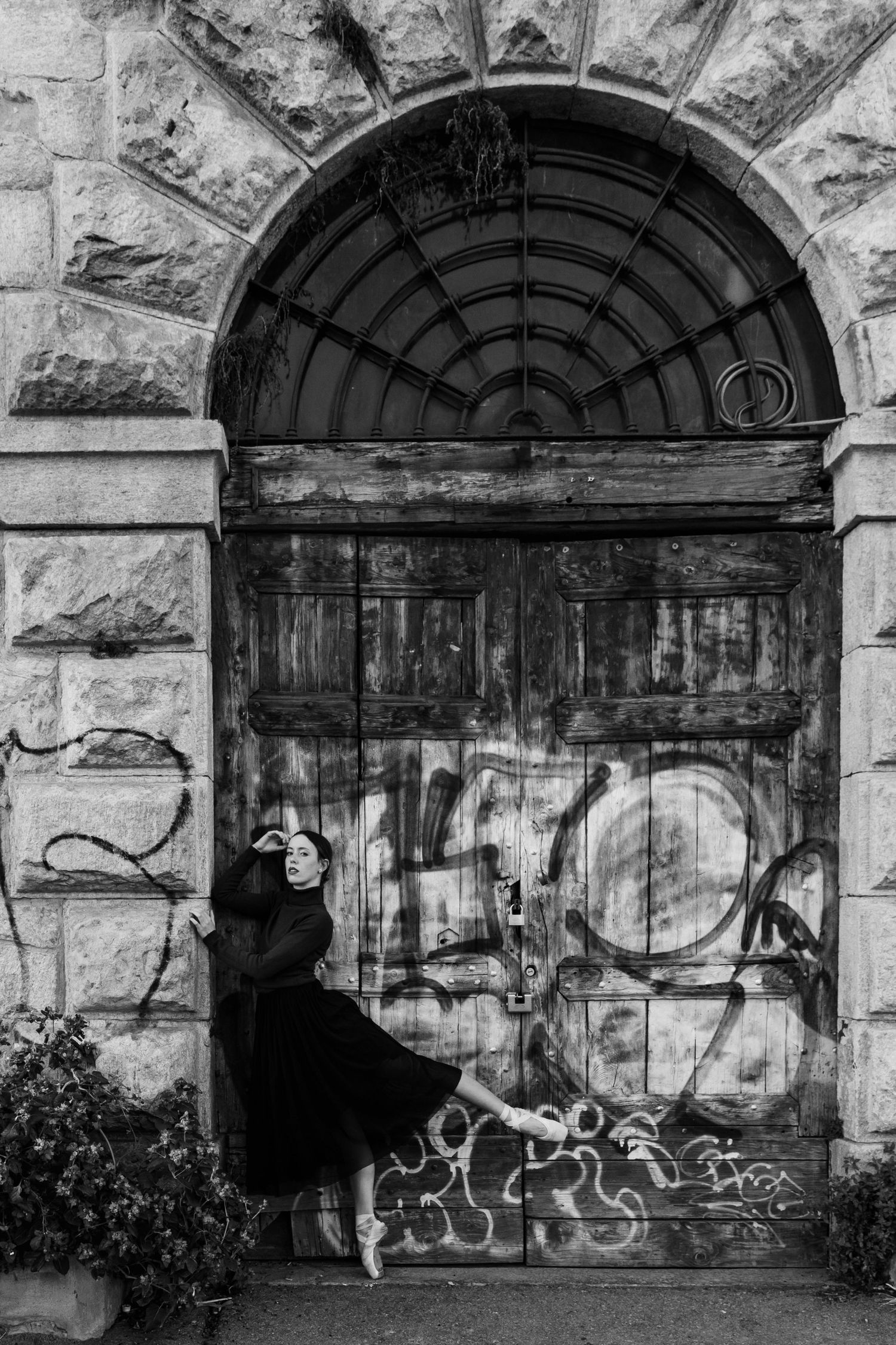 Ballet dancer in black dress posing en pointe before ornate graffiti-covered wooden door, black and white.