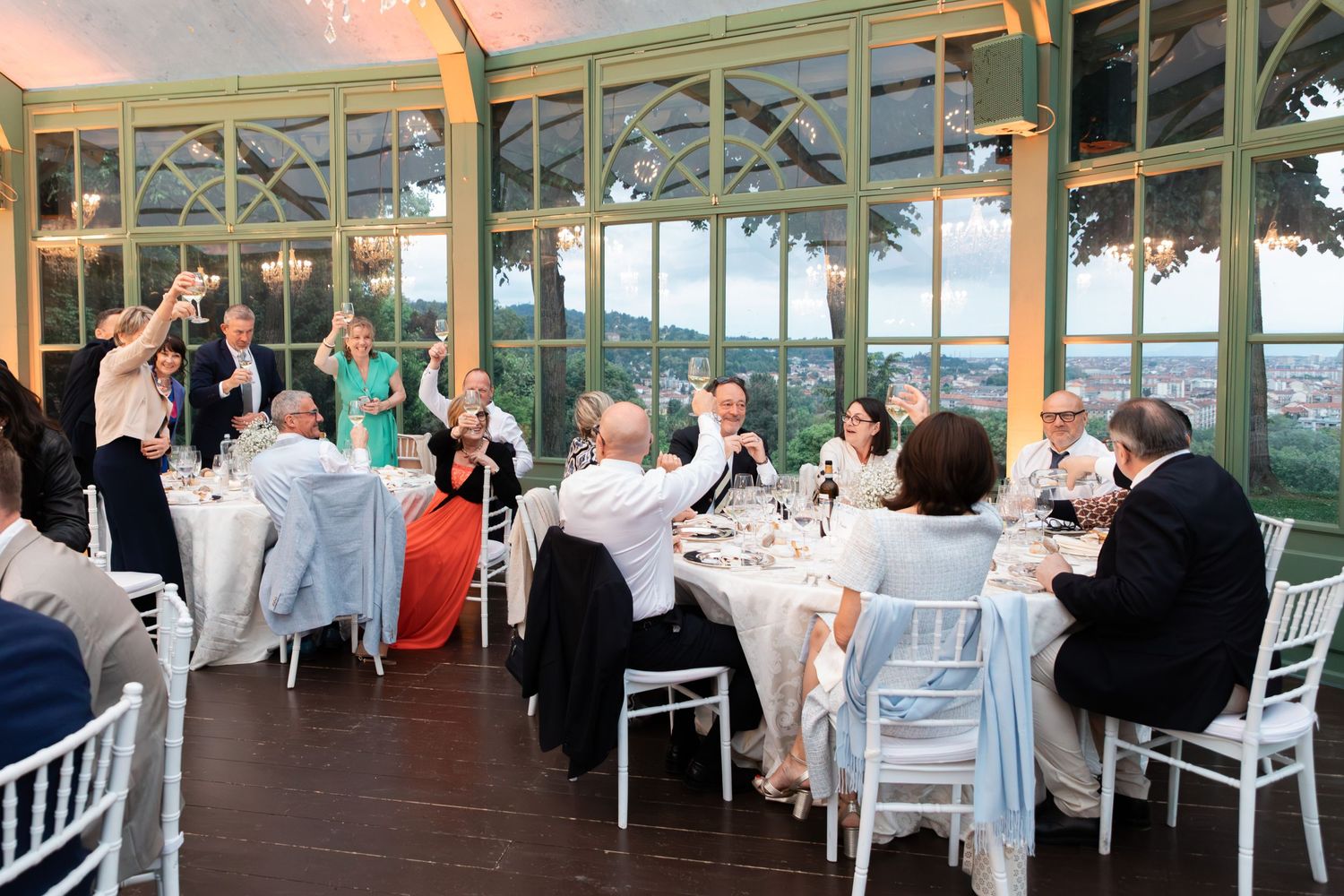 Wedding guests raising glasses in toast inside elegant glass conservatory with scenic view.