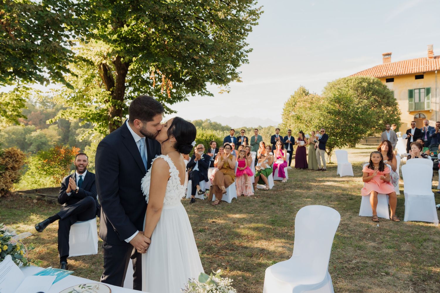 Bride and groom share first kiss at outdoor wedding ceremony with seated guests on sunny day.
