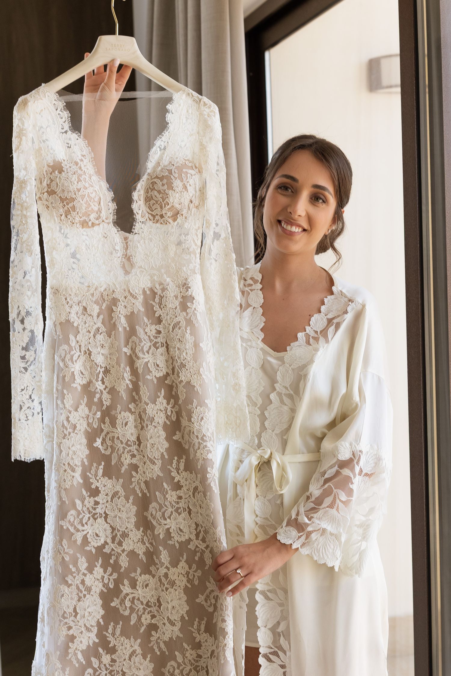 Smiling bride in white lace robe standing beside her elegant lace wedding dress hanging by a window.