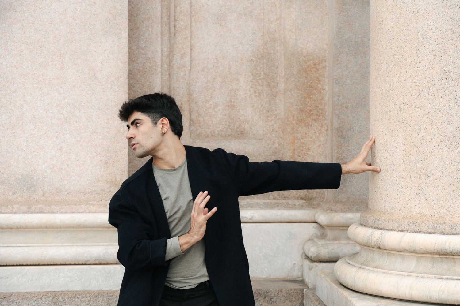 Male dancer in black coat performing expressive contemporary dance pose by stone columns.