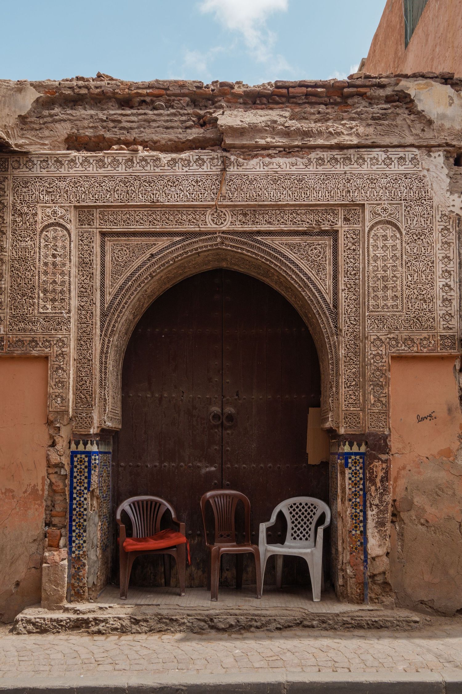 Elaborately carved Moroccan archway above a wooden door with plastic chairs on a cobblestone street.