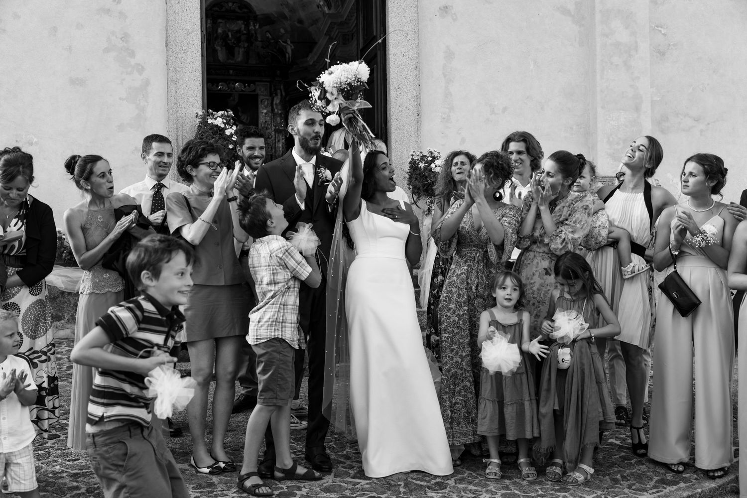 Newlyweds celebrate joyfully outside church surrounded by cheering wedding guests and children.
