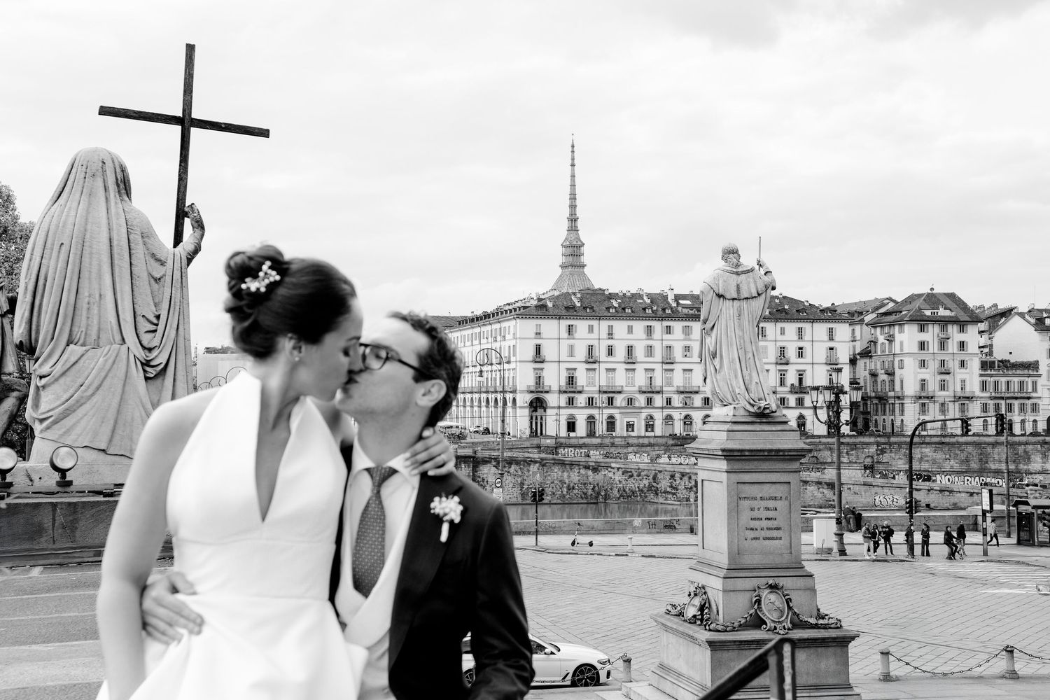 Black and white photo of couple kissing near statues with Turin cityscape in background.