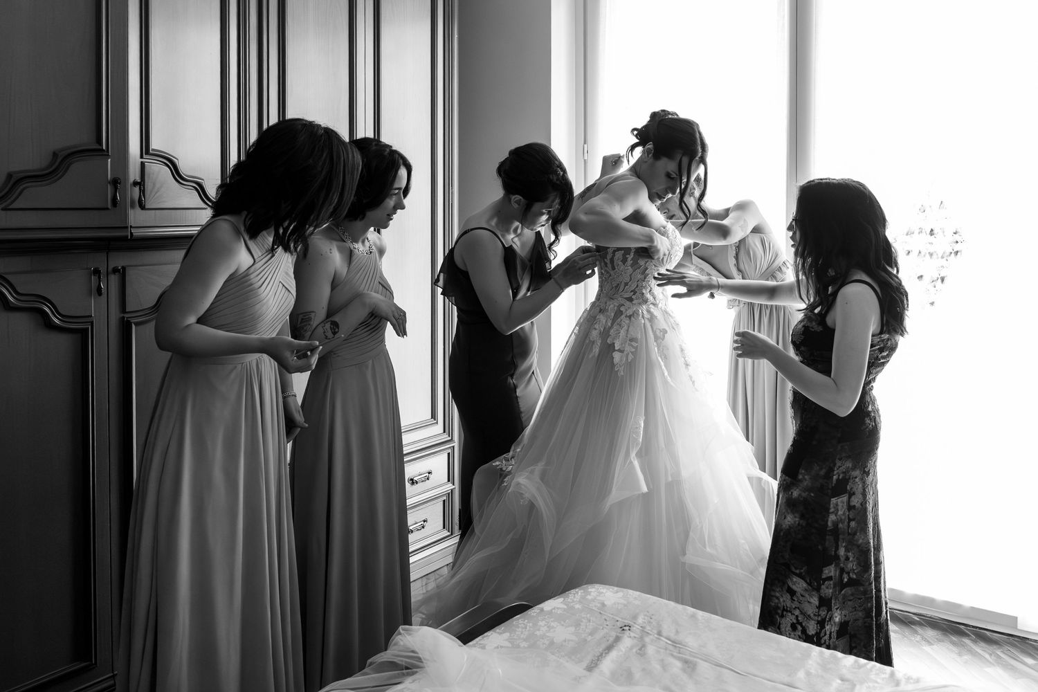 Black and white photo of bridesmaids helping bride into wedding dress near a window before ceremony.