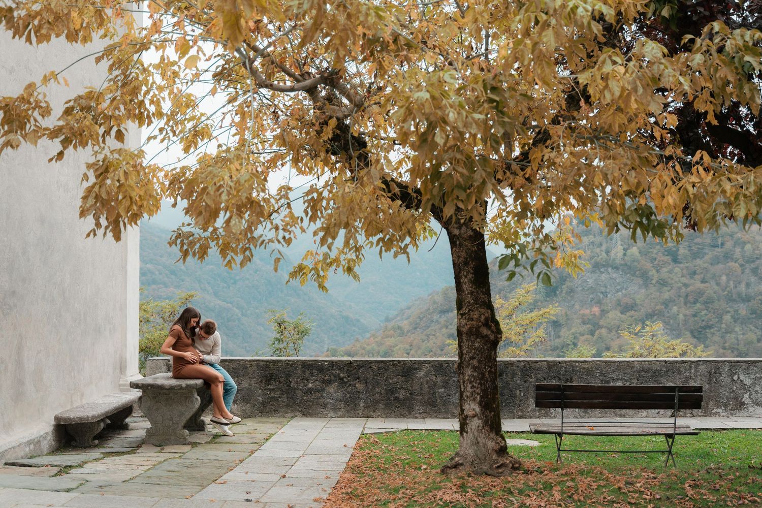 Two people sit closely together under an autumn tree with golden leaves, overlooking a scenic mountain valley.