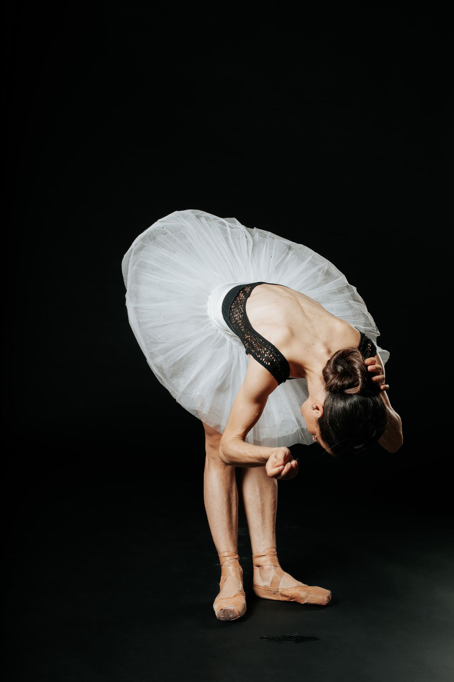 Ballerina in white tutu and black leotard bends forward gracefully against a dark black background.