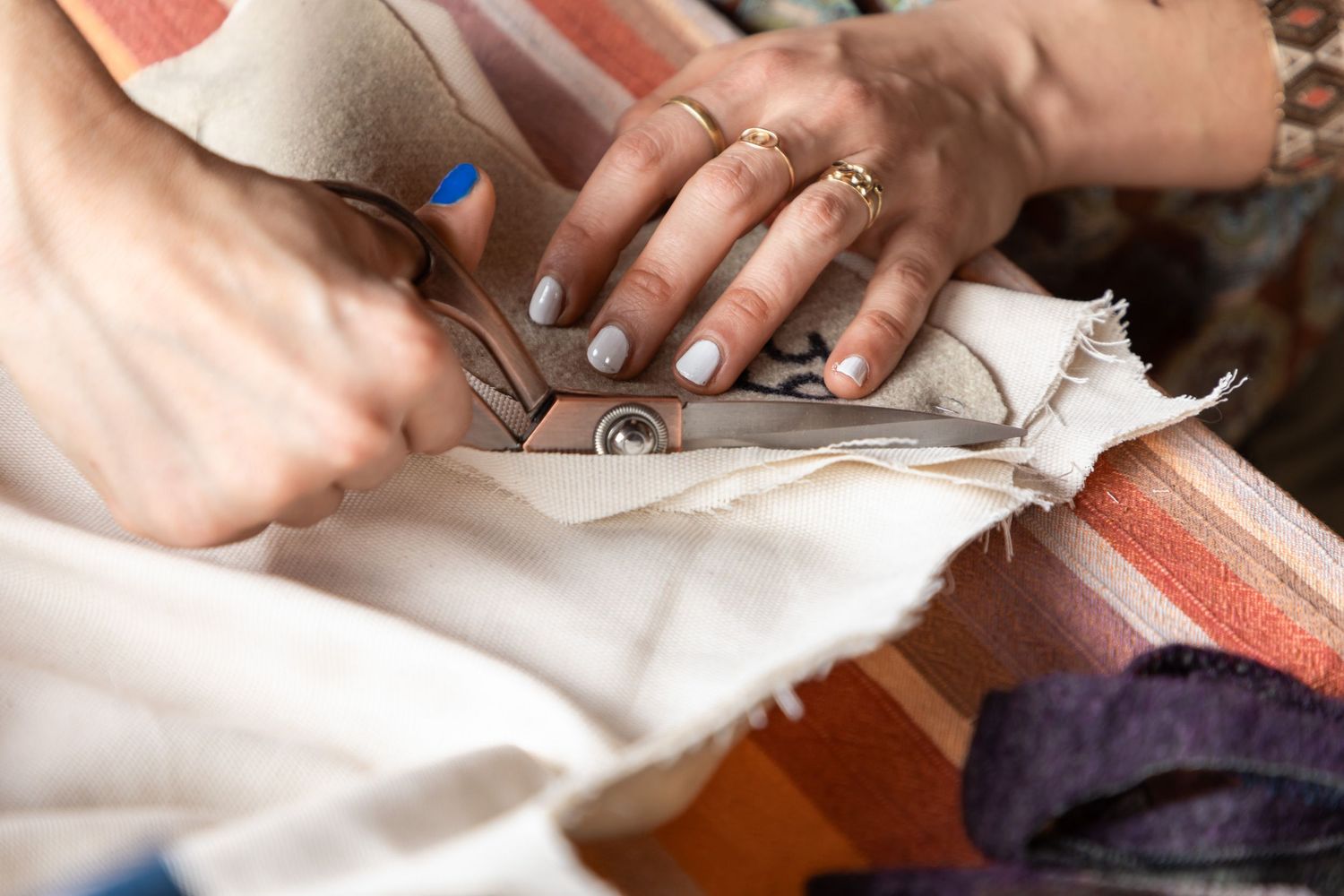 Hands with blue nails using scissors to cut white fabric on a striped surface.